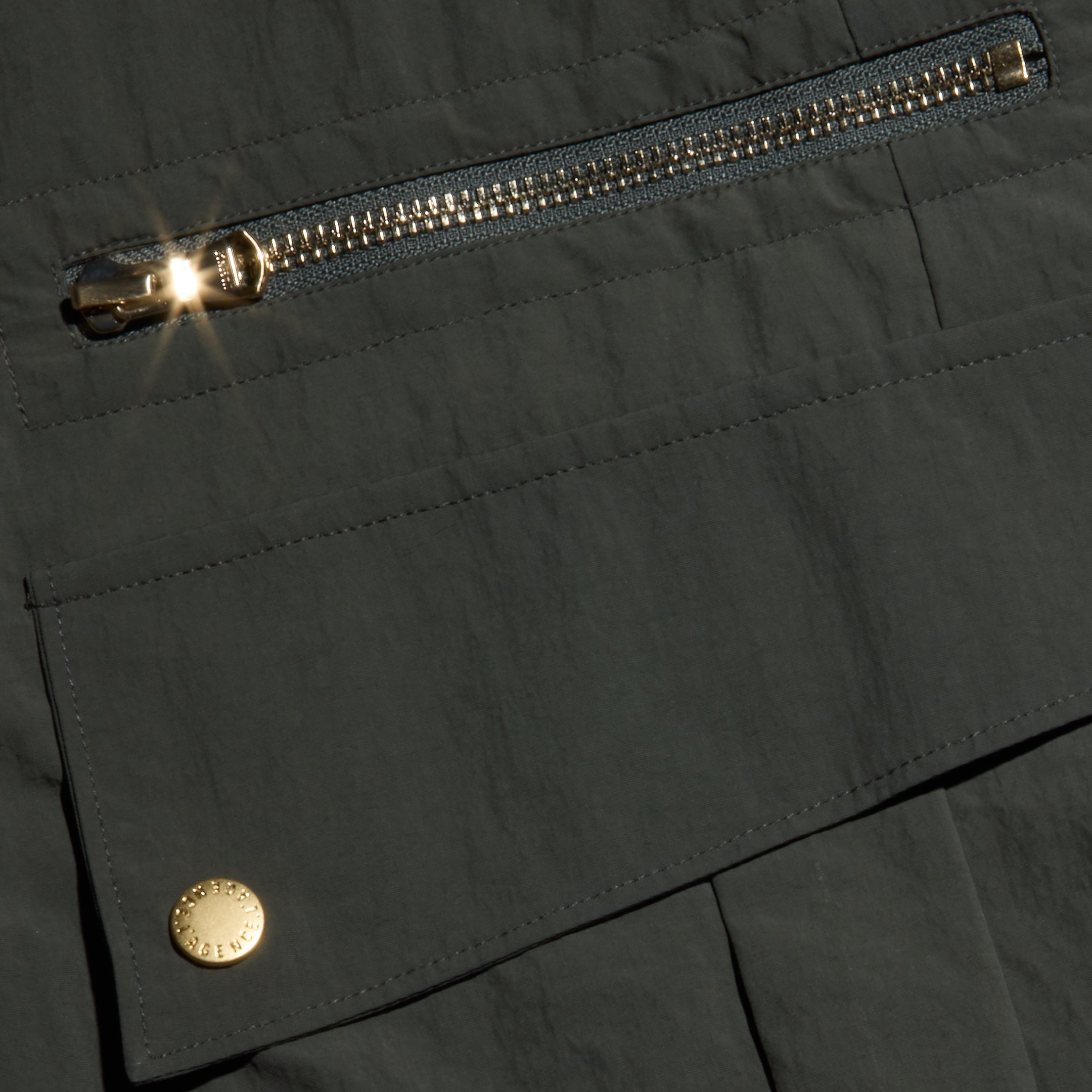 Close-up of the Greer Nylon Jogger in dark ash olive green, featuring a shiny metal zipper and snap button on a flap pocket—both catching the light. The design includes hints of cargo pockets that reflect its modern jogger style.