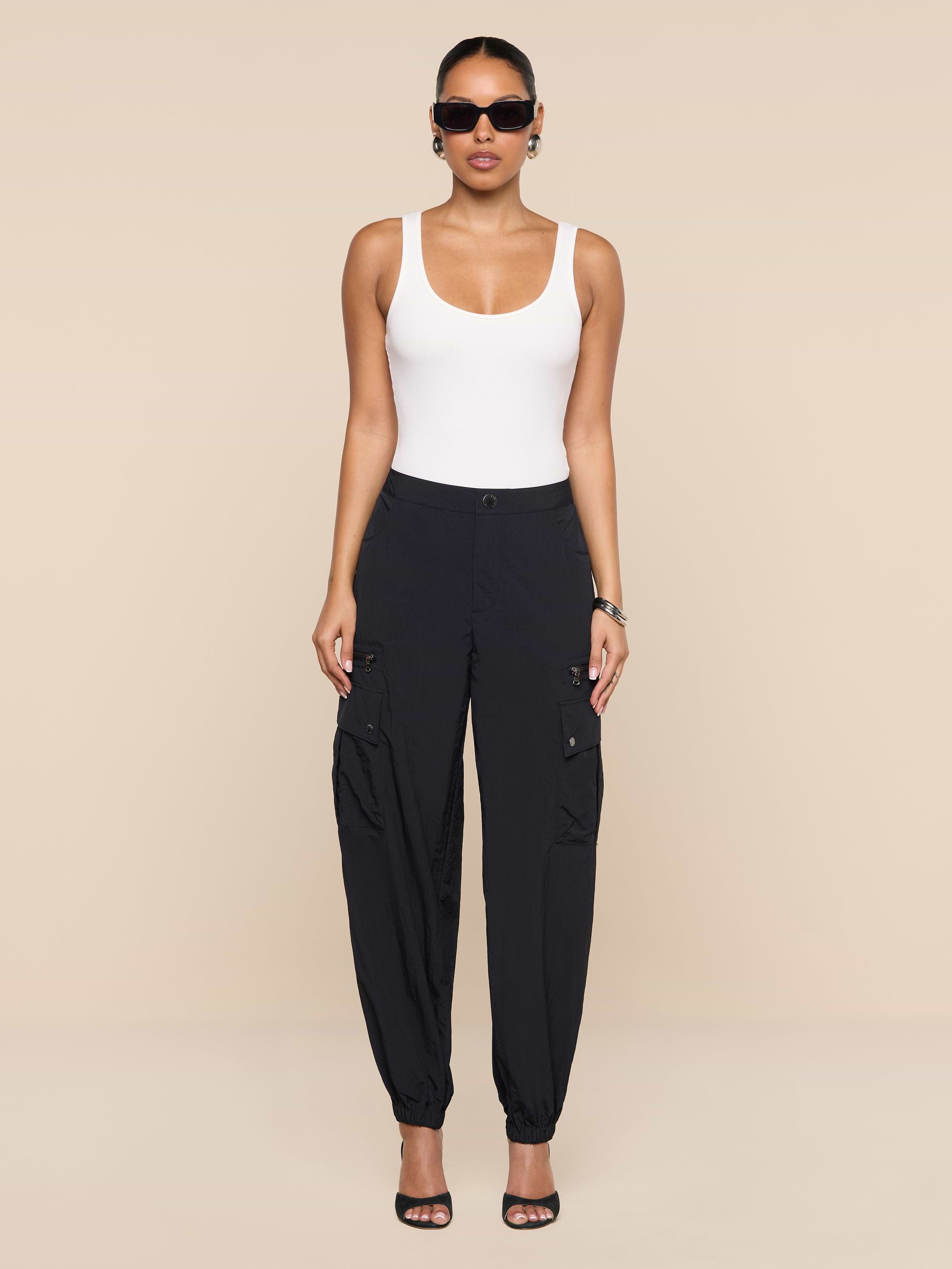 A woman wearing sunglasses, a white tank top, the Greer Nylon Jogger with cargo pockets in black, and black heeled sandals stands against a plain white background.