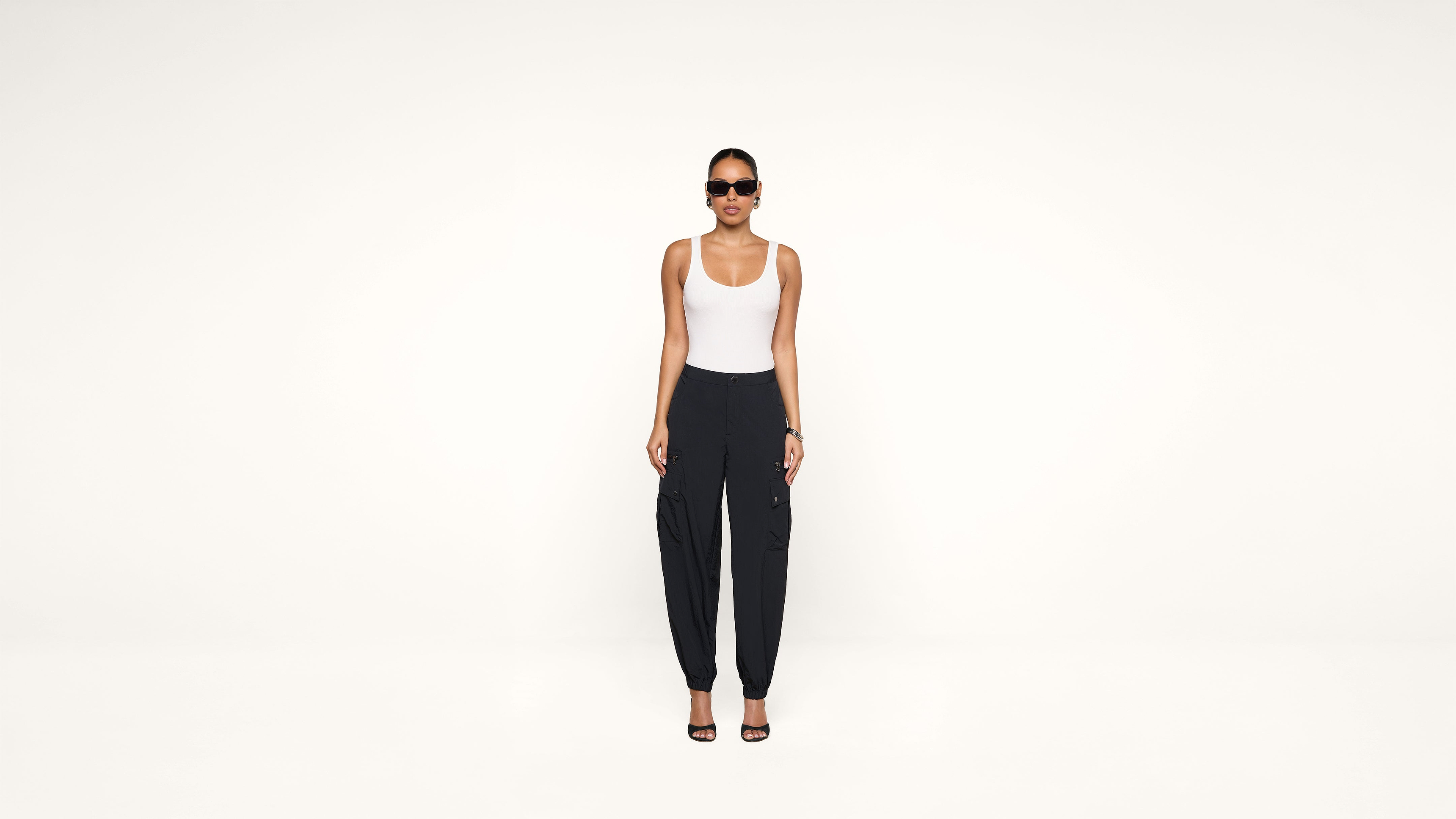 A woman wearing sunglasses, a white tank top, black cargo pants, and black heels stands facing forward against a plain white background.