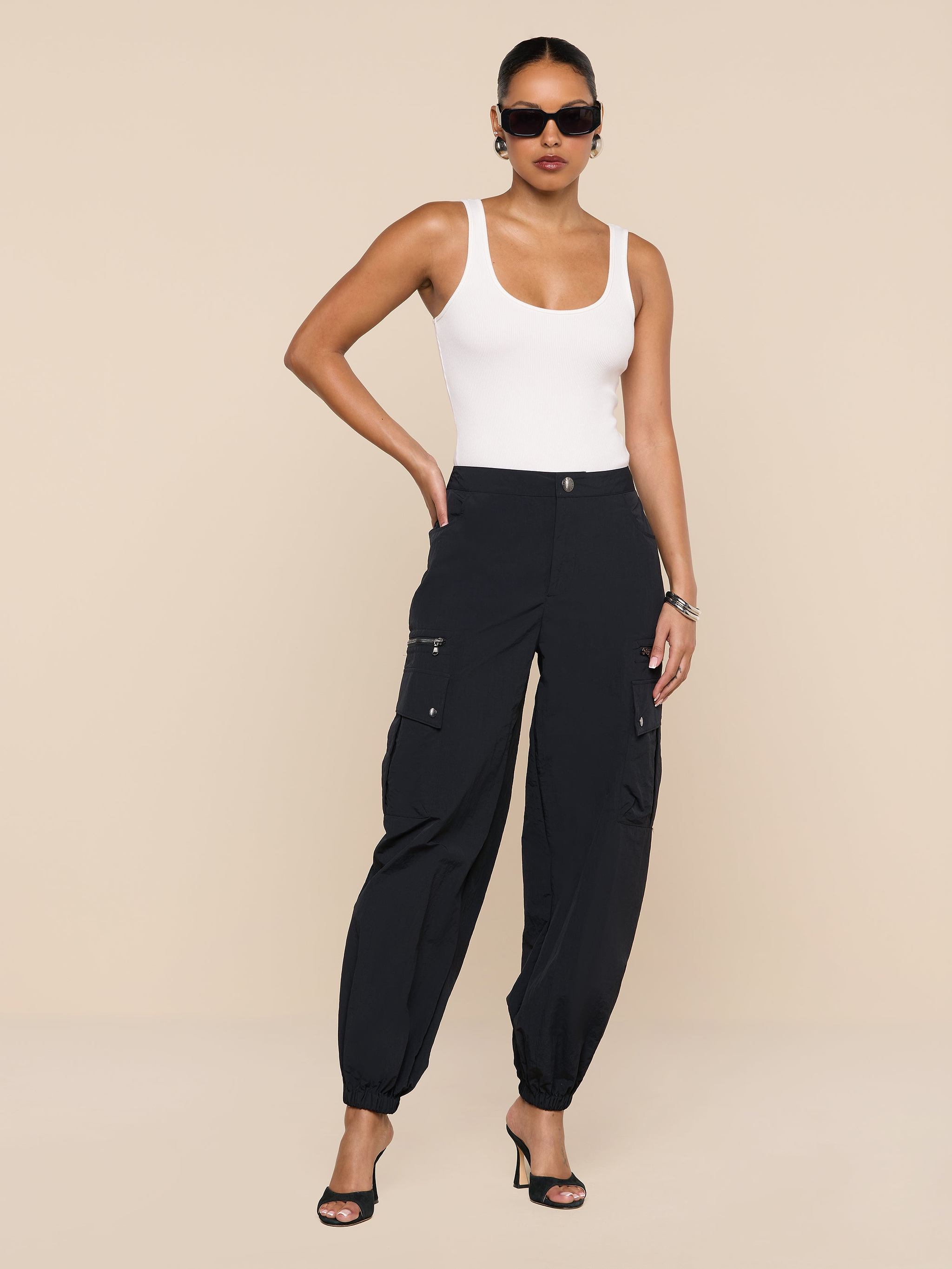 A woman stands against a plain white background, wearing the Greer Nylon Jogger with cargo pockets, a white tank top, black heeled sandals, large black sunglasses, hoop earrings, and her hair pulled back.