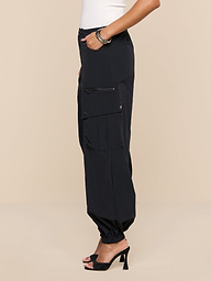 A person stands in profile wearing the Greer Nylon Jogger—black, water-repellent cargo jogger pants with cargo pockets—paired with a white top, black high heel sandals, and bracelets. The background is plain white.