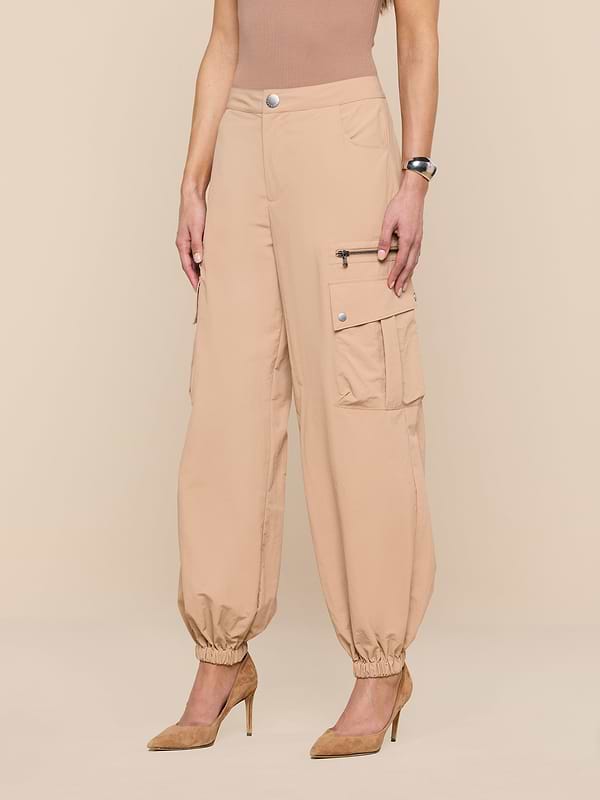 A woman wears Greer Nylon Jogger cargo pants with elastic cuffs, cargo pockets, and zipper detail, paired with a sleeveless taupe top and tan pointed-toe heels. Her left wrist shows a watch as she stands with arms at her sides.