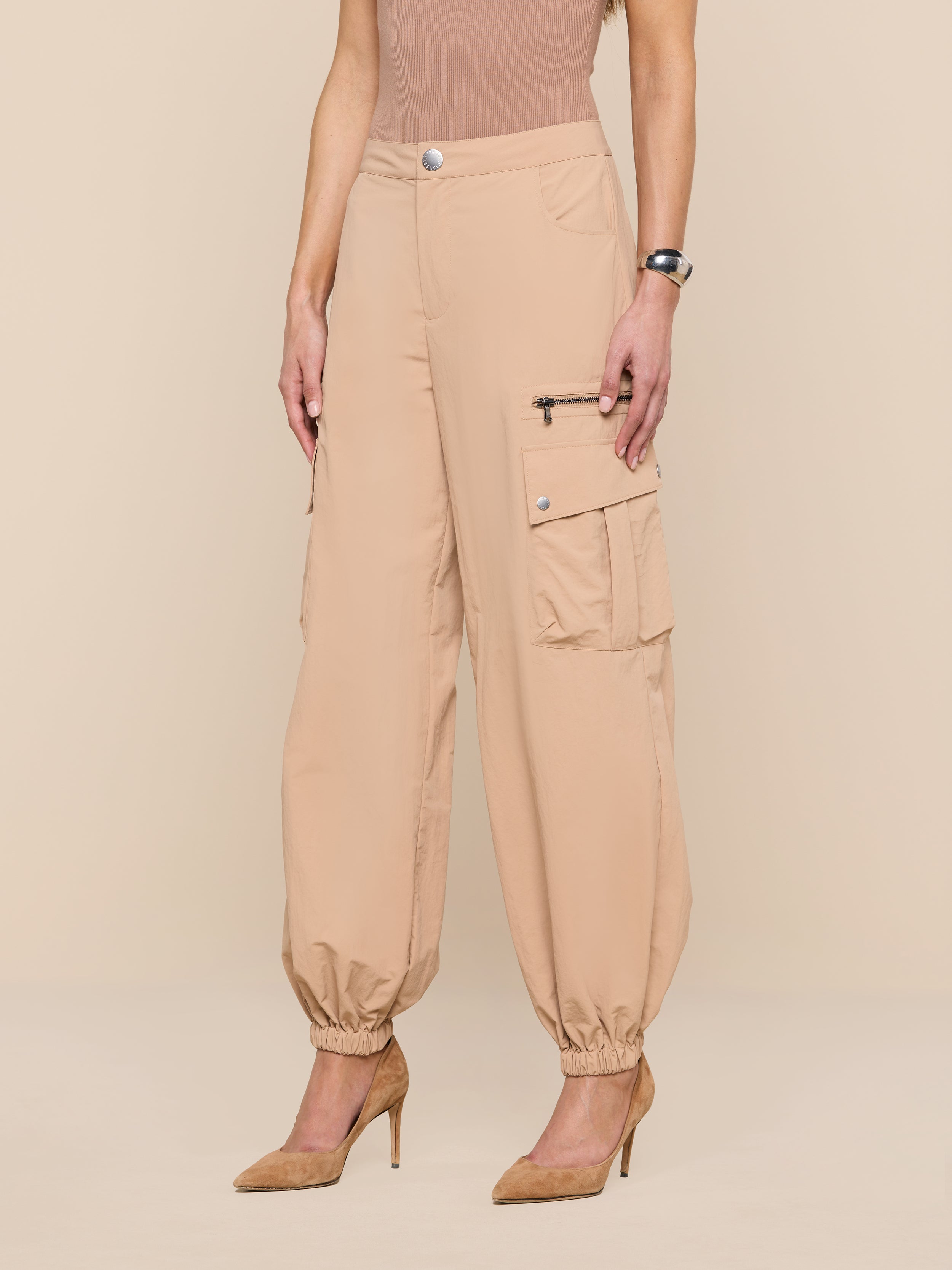 A woman wears Greer Nylon Jogger cargo pants with elastic cuffs, cargo pockets, and zipper detail, paired with a sleeveless taupe top and tan pointed-toe heels. Her left wrist shows a watch as she stands with arms at her sides.