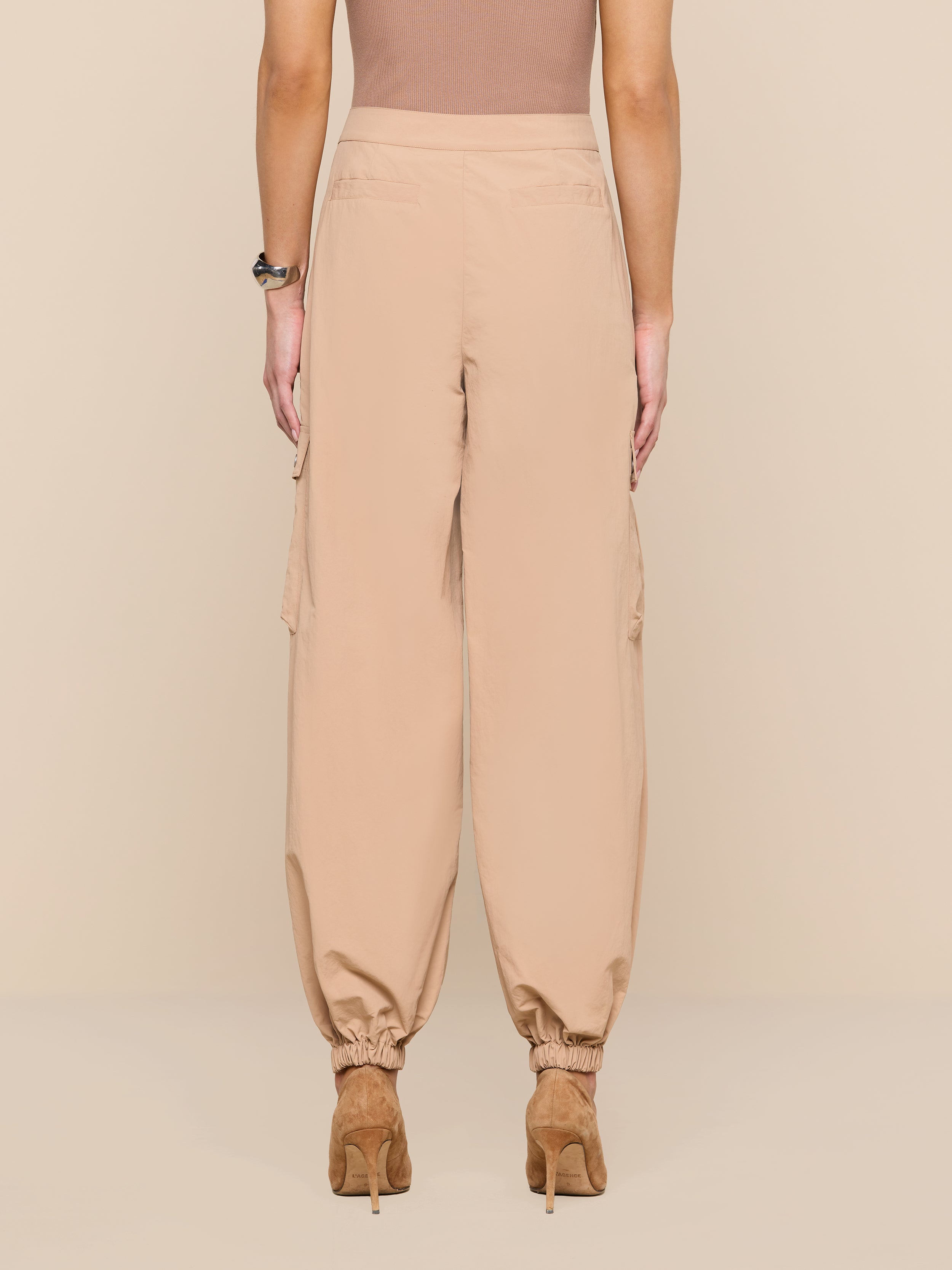 A person is shown from the back against a plain beige background wearing the Greer Nylon Jogger—modern, high-waisted beige cargo pants with elastic cuffs, cargo pockets—and tan heeled shoes.
