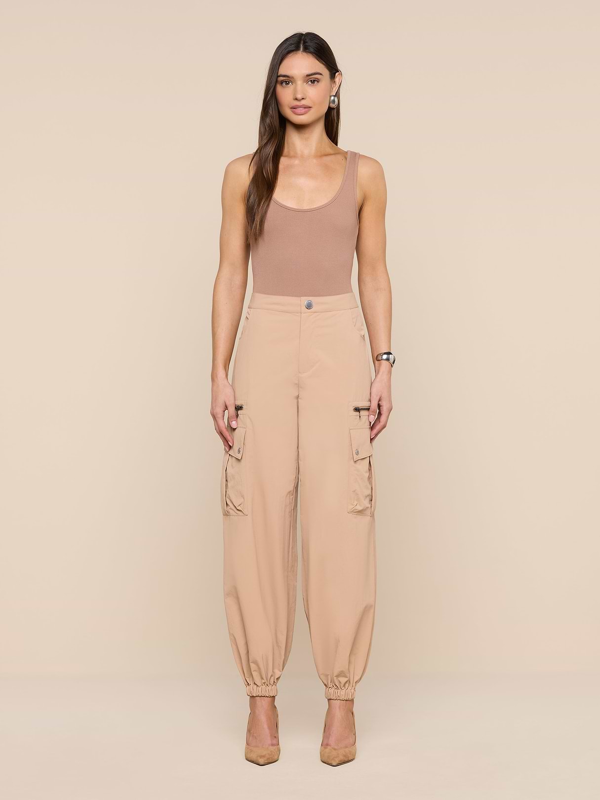 A woman stands facing forward against a plain white background, wearing a tan sleeveless top, the Greer Nylon Jogger—high-waisted beige cargo pants with cargo pockets—and beige heels.