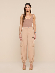 A woman with long brown hair stands against a beige background wearing the Greer Nylon Jogger, a sleeveless tan top, and beige heels. The joggers feature roomy cargo pockets. She looks straight ahead with arms relaxed at her sides.