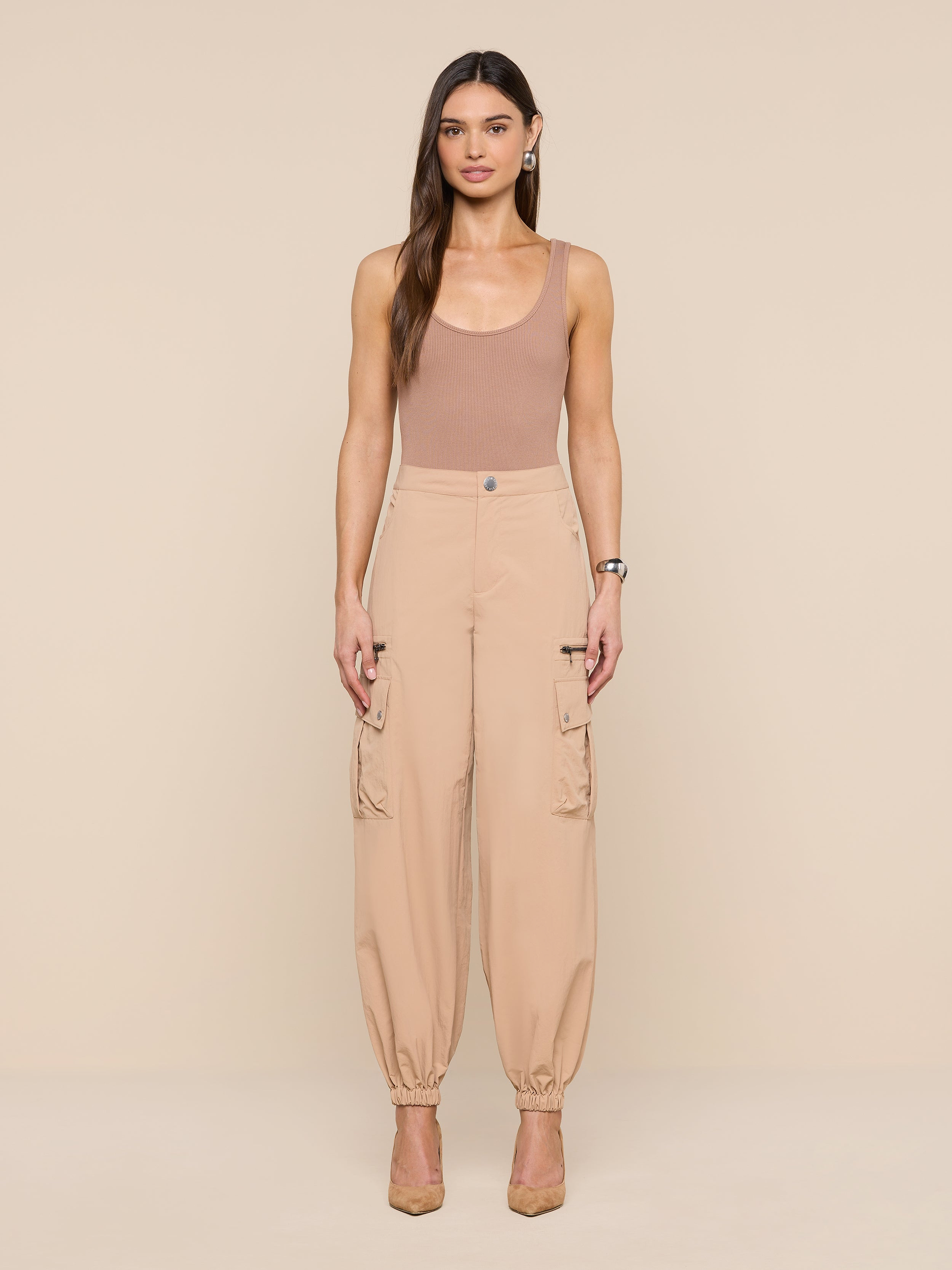 A woman with long brown hair stands against a beige background wearing the Greer Nylon Jogger, a sleeveless tan top, and beige heels. The joggers feature roomy cargo pockets. She looks straight ahead with arms relaxed at her sides.