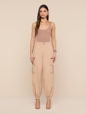A woman with long brown hair stands against a beige background wearing the Greer Nylon Jogger, a sleeveless tan top, and beige heels. The joggers feature roomy cargo pockets. She looks straight ahead with arms relaxed at her sides.