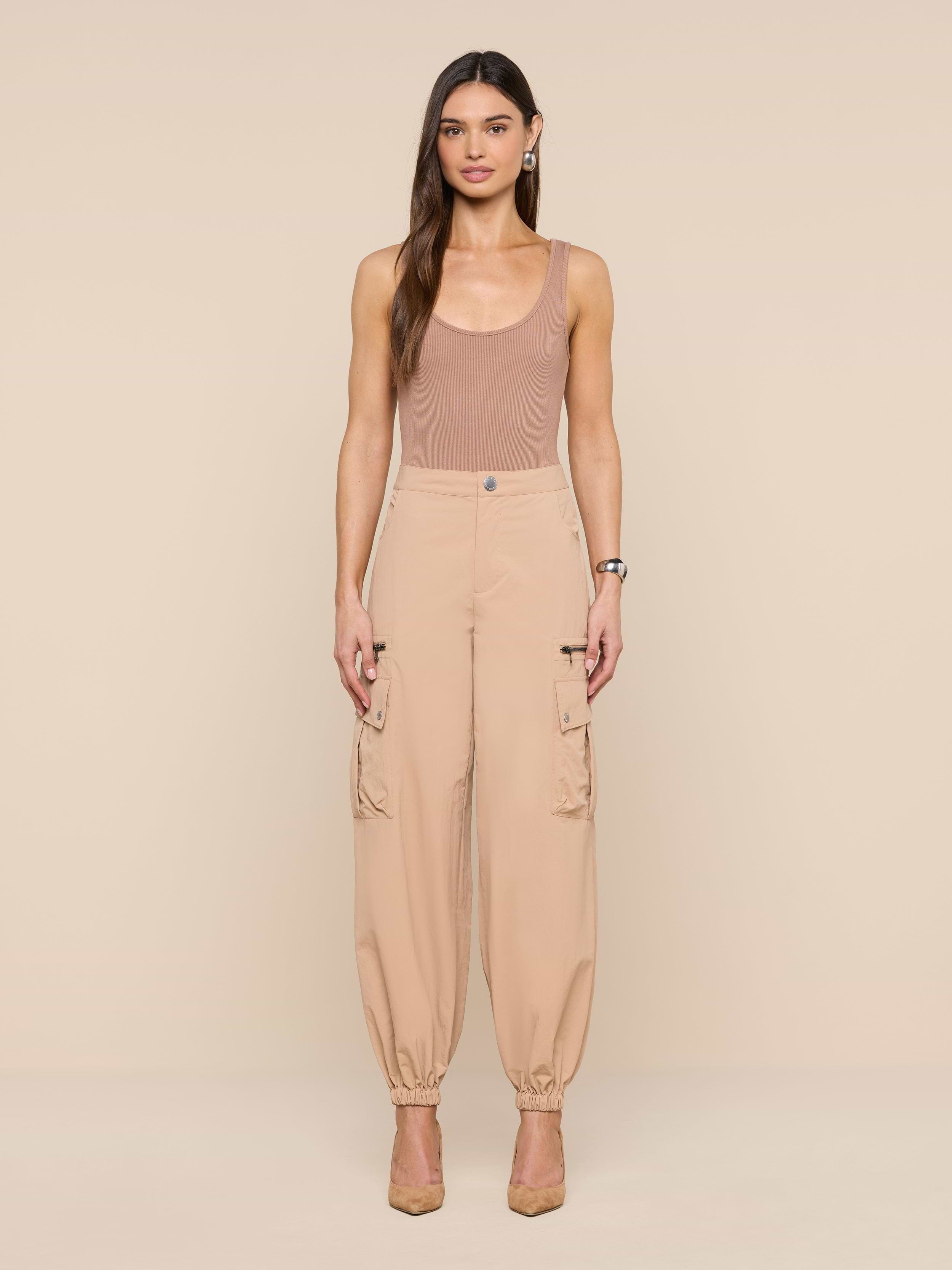 A woman with long brown hair stands against a beige background wearing the Greer Nylon Jogger, a sleeveless tan top, and beige heels. The joggers feature roomy cargo pockets. She looks straight ahead with arms relaxed at her sides.