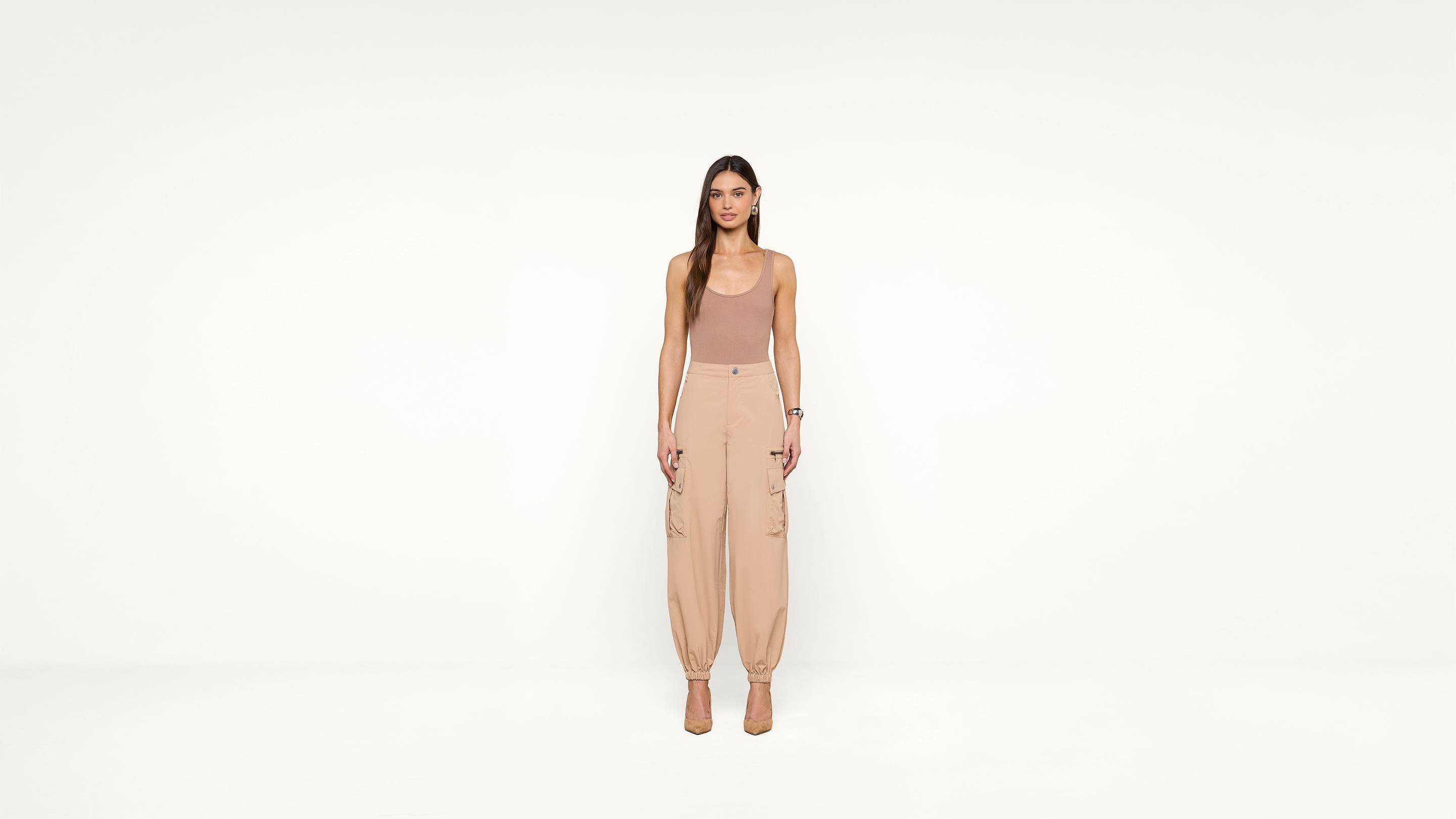 A woman stands facing forward in a neutral-toned outfit, including a sleeveless top and high-waisted pants, against a plain white background.
