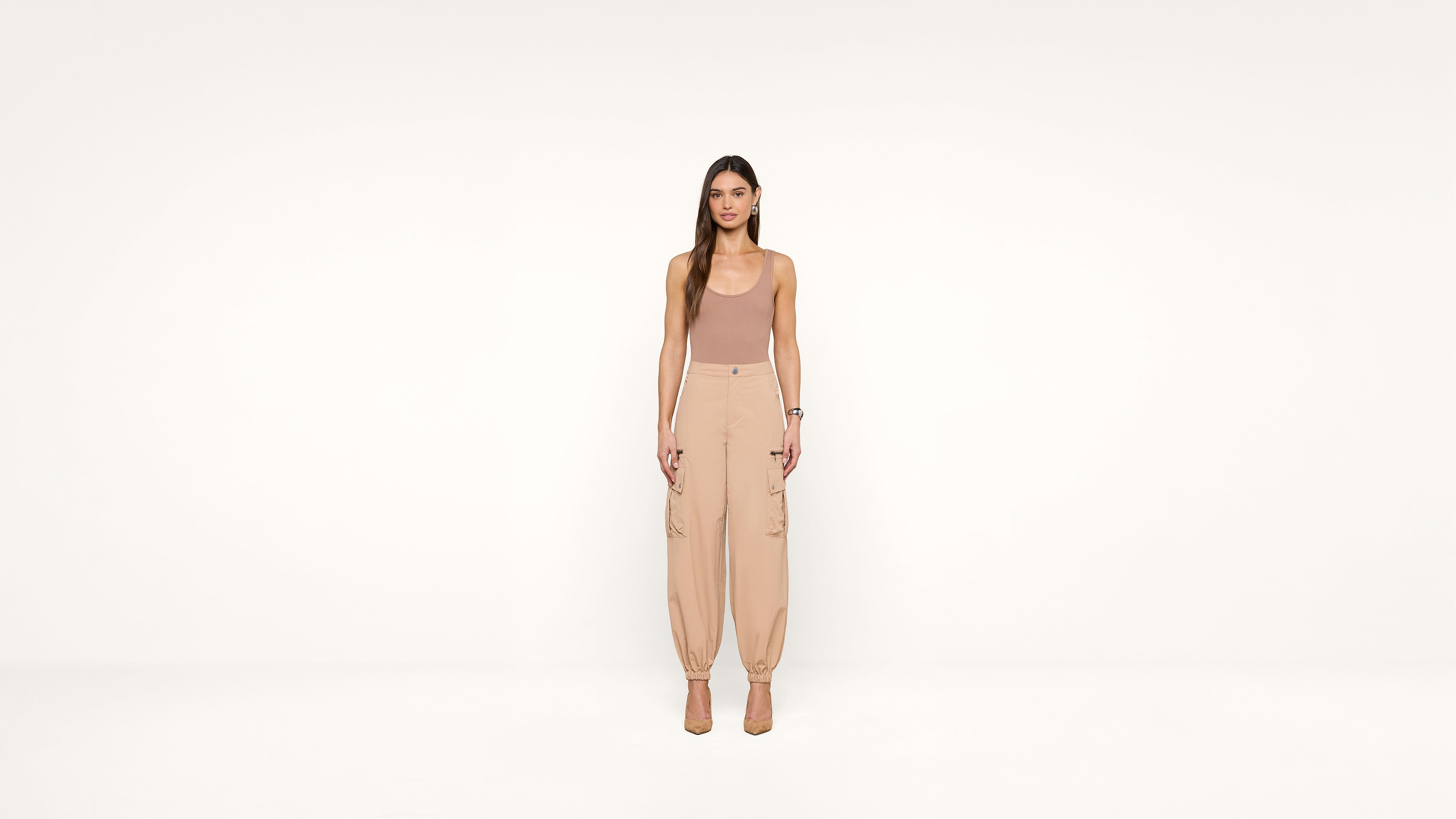 A woman stands facing forward in a neutral-toned outfit, including a sleeveless top and high-waisted pants, against a plain white background.