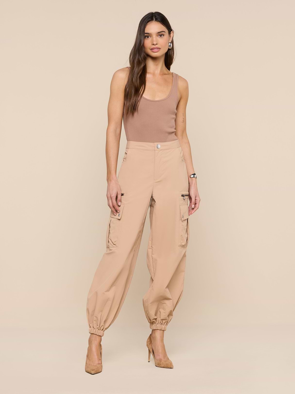 A woman stands against a white background wearing the Greer Nylon Jogger, a pair of beige jogger-style cargo pants with cargo pockets, paired with a sleeveless beige top and beige high heels. Her hair is down, and she has a neutral expression.
