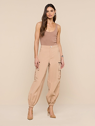 A woman with long, wavy hair and a neutral expression models the Greer Nylon Jogger—high-waisted tan cargo pants—paired with a brown tank top and tan heels against a beige background, highlighting modern jogger style.