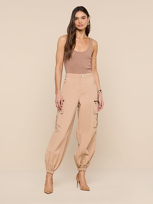 A woman with long, wavy hair and a neutral expression models the Greer Nylon Jogger—high-waisted tan cargo pants—paired with a brown tank top and tan heels against a beige background, highlighting modern jogger style.