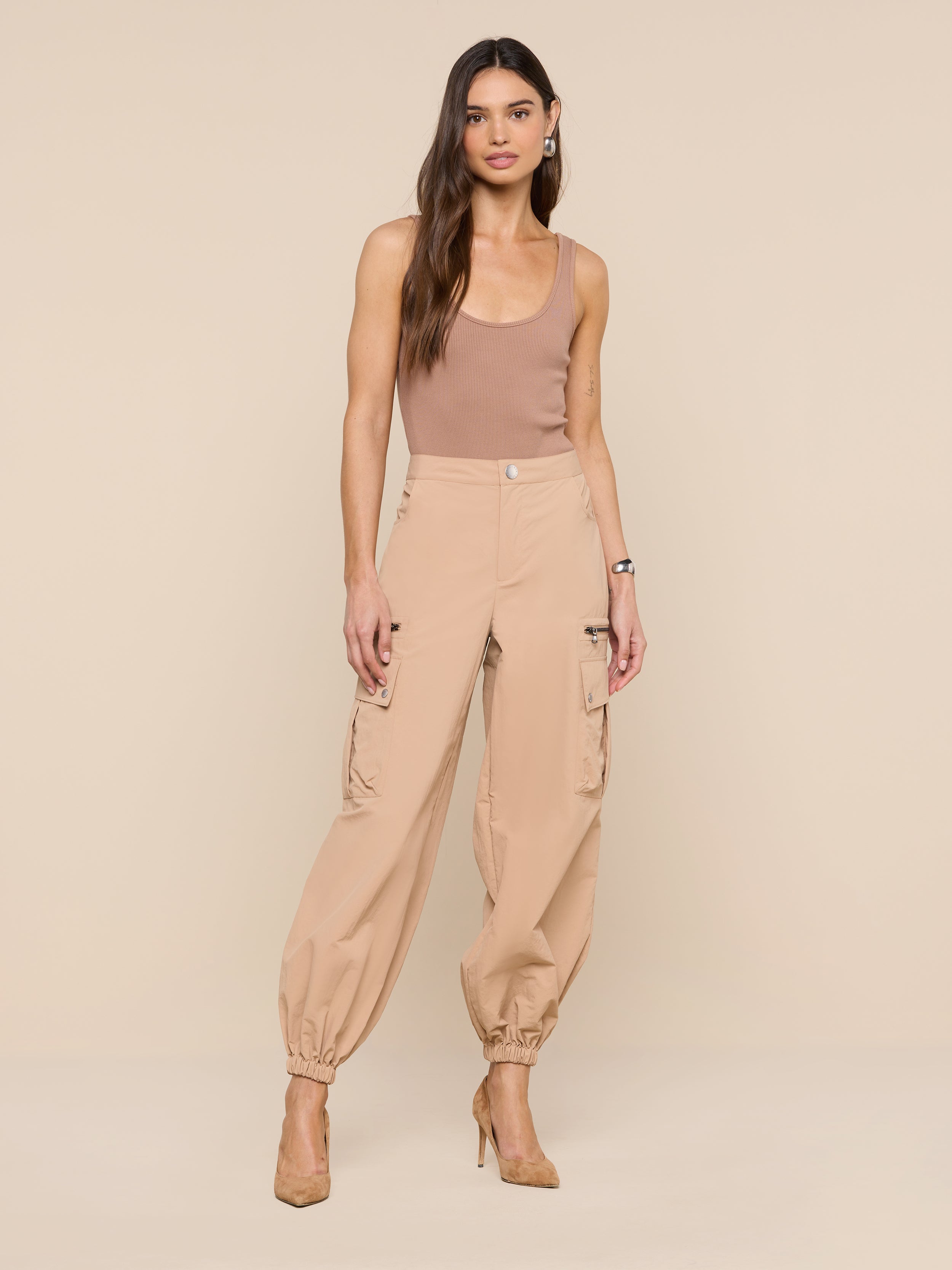 A woman with long, wavy hair and a neutral expression models the Greer Nylon Jogger—high-waisted tan cargo pants—paired with a brown tank top and tan heels against a beige background, highlighting modern jogger style.