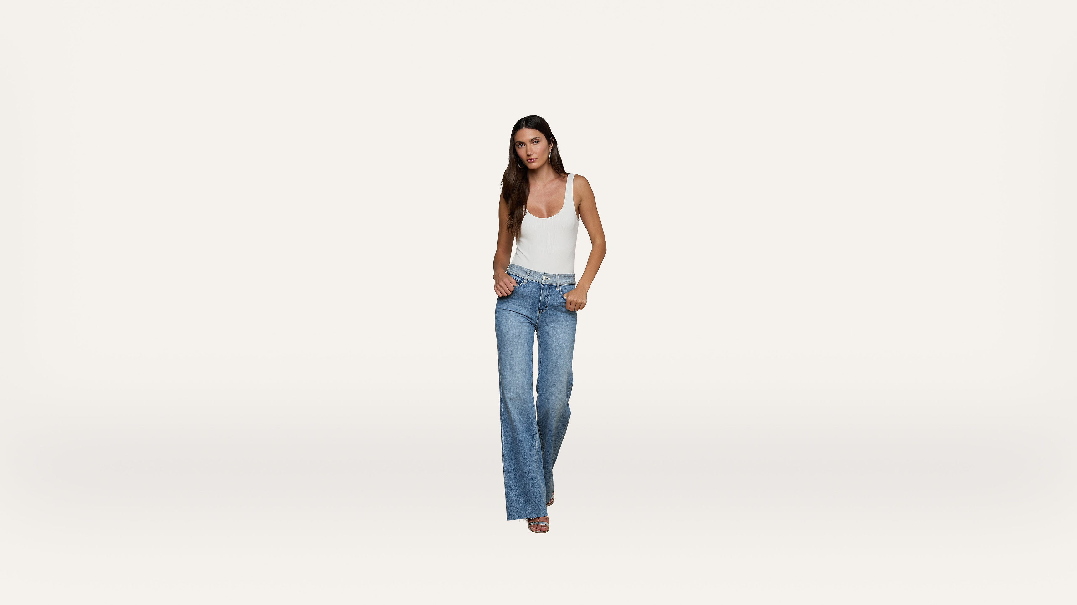 Woman with long hair wears a white tank top and high-waisted blue jeans, standing against a plain white background.
