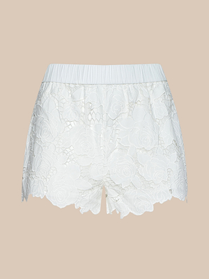 The Laina Eyelet Short features white floral lace, cotton eyelet detailing, scalloped hem, lined pockets, and an elastic waistband, shown against a plain beige background.