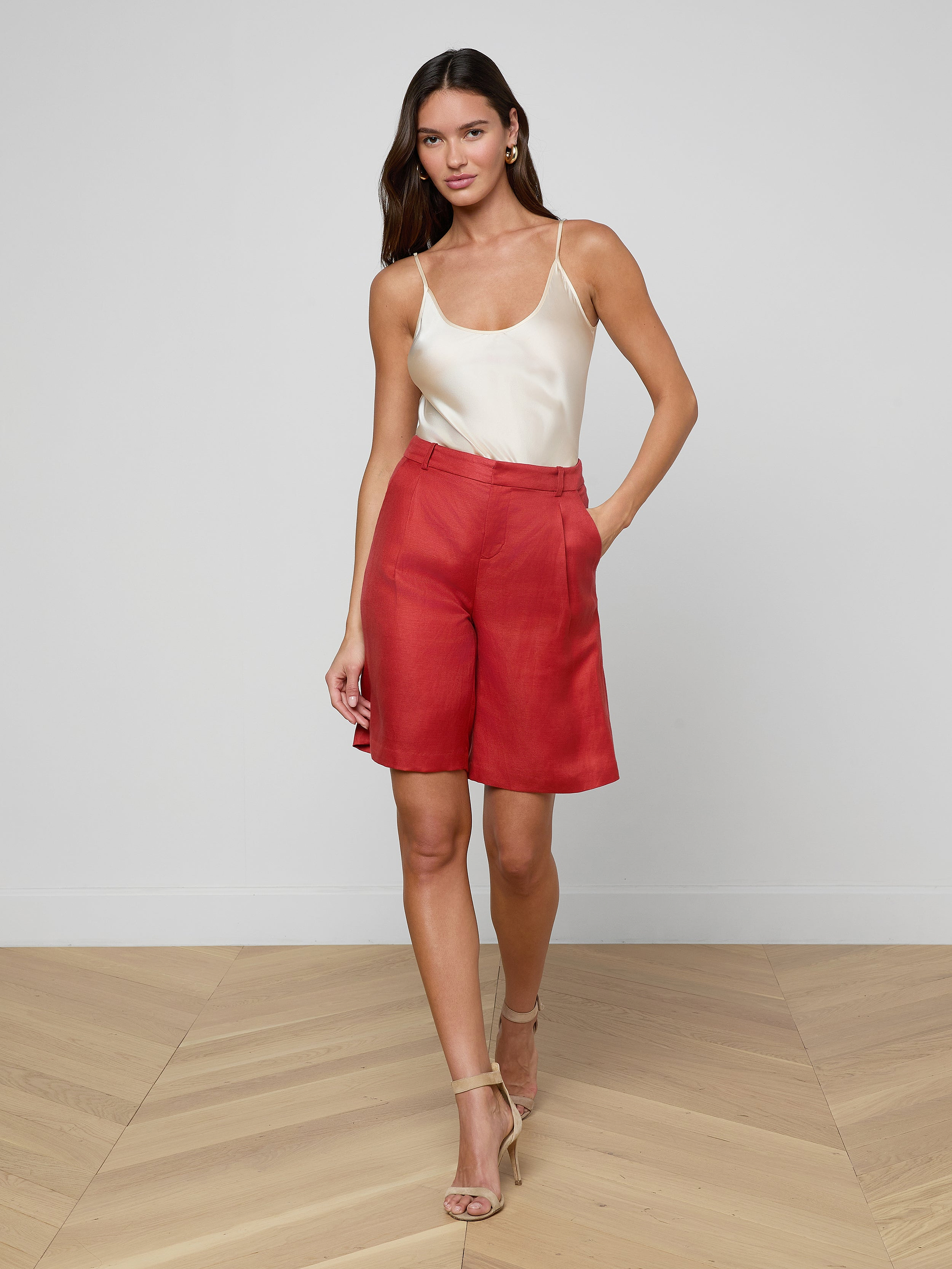 A woman stands against a plain white background with one hand in her pocket, wearing a white sleeveless top, beige high-heeled sandals, and the Doretta Bermuda Short in red—a perfect look for summer-in-the-office style.