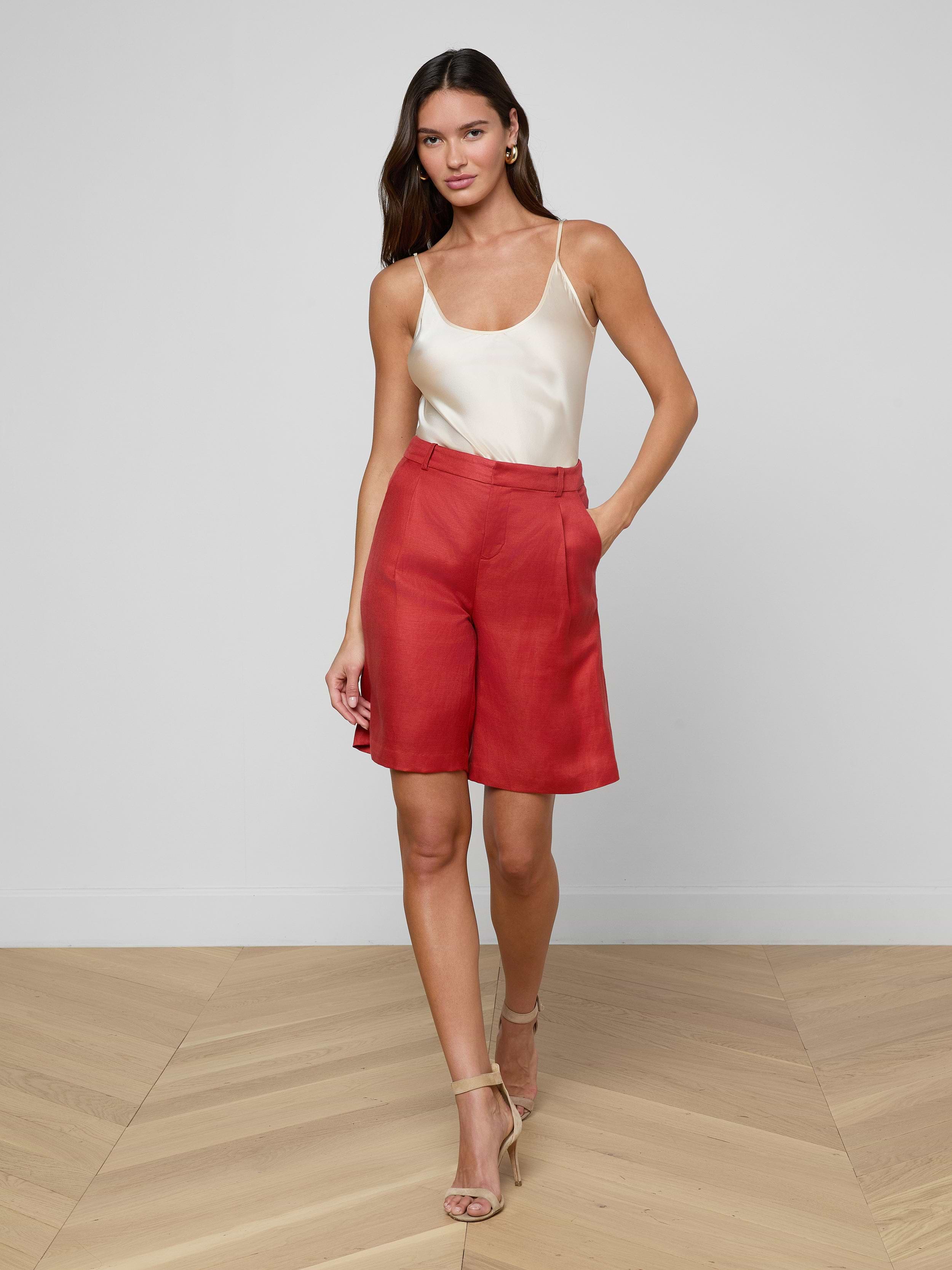 A woman stands against a plain white background with one hand in her pocket, wearing a white sleeveless top, beige high-heeled sandals, and the Doretta Bermuda Short in red—a perfect look for summer-in-the-office style.