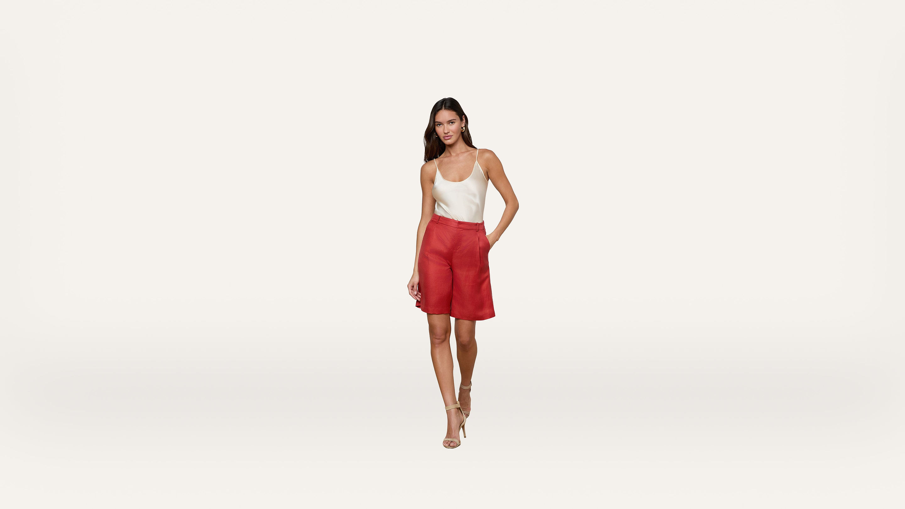 A woman stands against a plain white background, wearing a white sleeveless top, red skirt, and beige high-heeled shoes. She has one hand in her skirt pocket and is smiling slightly.