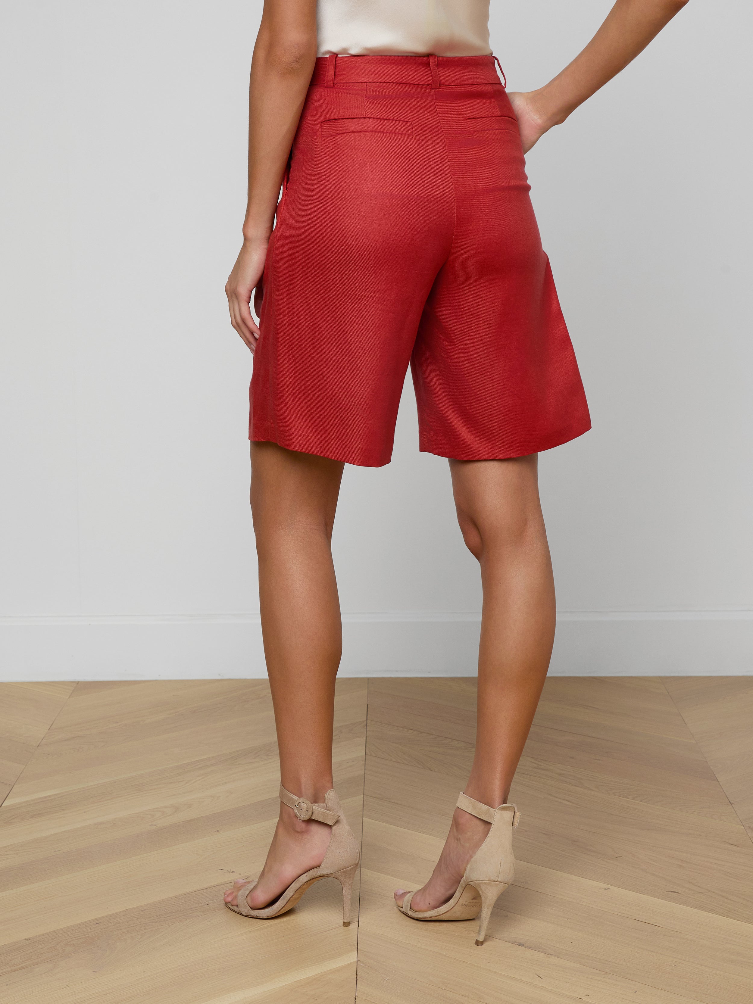 A woman stands with her back to the camera, wearing the Doretta Bermuda Short in a red linen blend, paired with a light-colored top and beige high-heeled sandals against a plain white background—perfect for summer-in-the-office style.