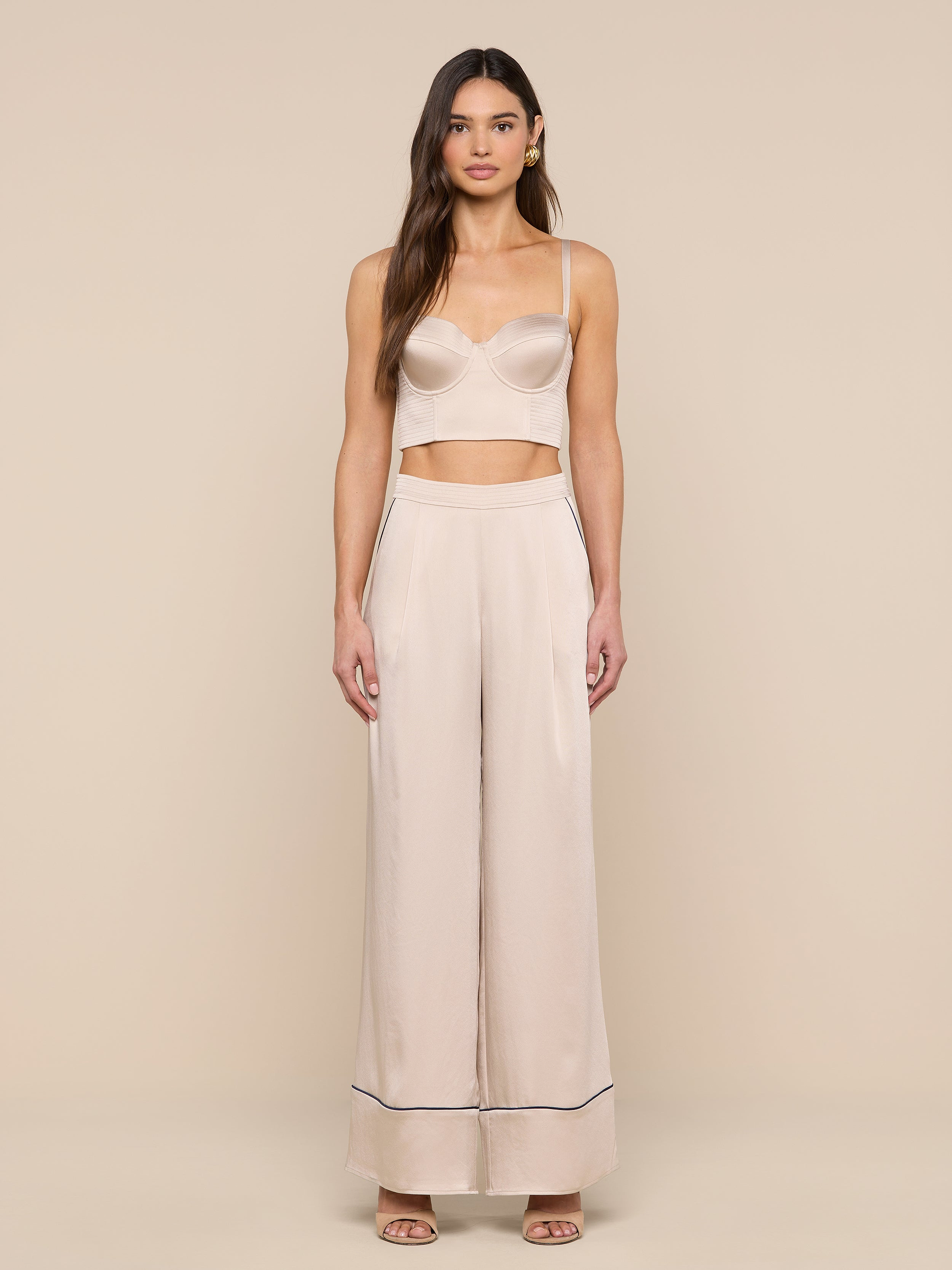 A woman stands facing forward wearing a light beige crop top and the Tavia Trouser — a high-waisted, wide-leg satin pant with dark trim at the hem — paired with open-toe shoes. The background is plain white.