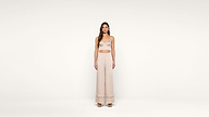 A woman stands facing forward in a minimalist setting, wearing a light beige crop top and matching wide-leg pants with sandals. The background is plain white.