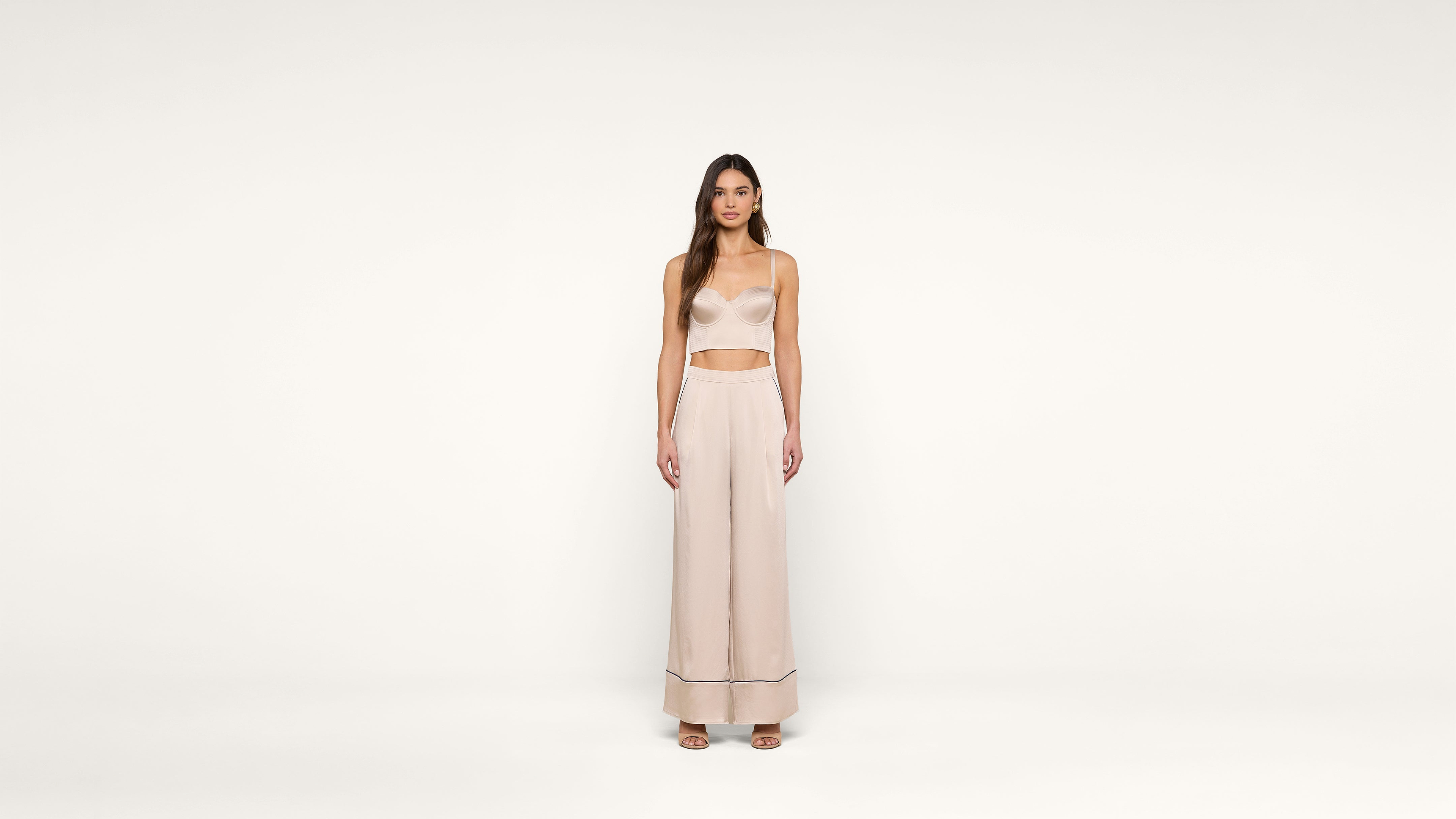 A woman stands facing forward in a minimalist setting, wearing a light beige crop top and matching wide-leg pants with sandals. The background is plain white.