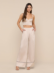 A woman stands against a plain white background wearing the Nysa Bustier Top—a cream-colored crop top styled for a supportive fit—and matching high-waisted wide-leg pants with side pockets and heeled sandals. She has long, loose hair and a neutral expression.