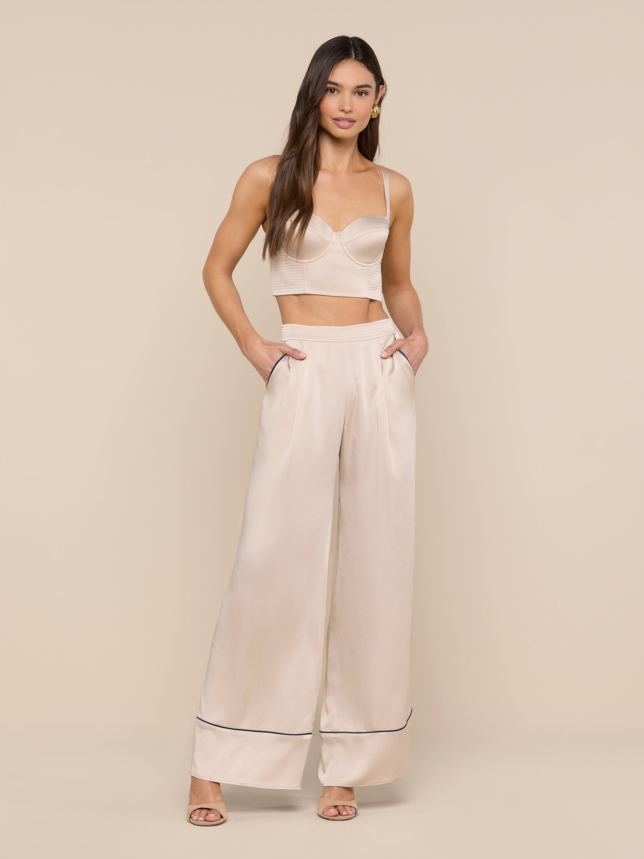 A woman stands against a plain white background wearing the Nysa Bustier Top—a cream-colored crop top styled for a supportive fit—and matching high-waisted wide-leg pants with side pockets and heeled sandals. She has long, loose hair and a neutral expression.