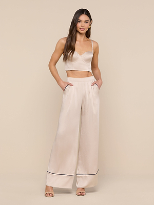 A woman poses against a beige background in luxe loungewear: a light satin crop top with thin straps, paired with the Tavia Trouser—high-waisted wide-leg pull-ons featuring black trim. Her long hair and open-toe heels complete the chic look.