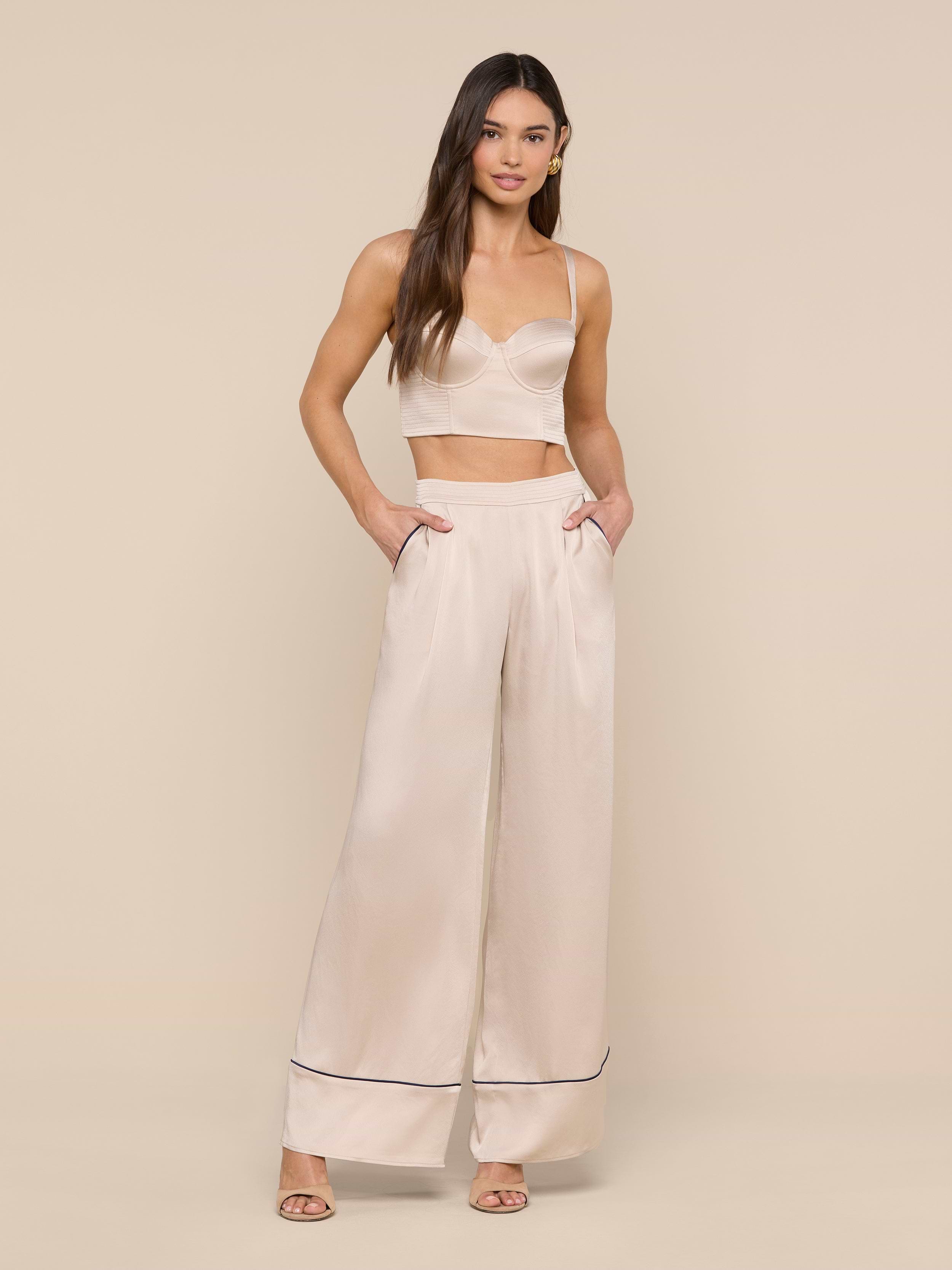 A woman stands against a plain white background wearing the Nysa Bustier Top—a cream-colored crop top styled for a supportive fit—and matching high-waisted wide-leg pants with side pockets and heeled sandals. She has long, loose hair and a neutral expression.