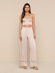 A woman stands against a plain white background wearing a light beige camisole and the Tavia Trouser in matching satin with dark trim at the hems. She has long brown hair and is wearing open-toe heels, capturing the effortless elegance of luxe loungewear.