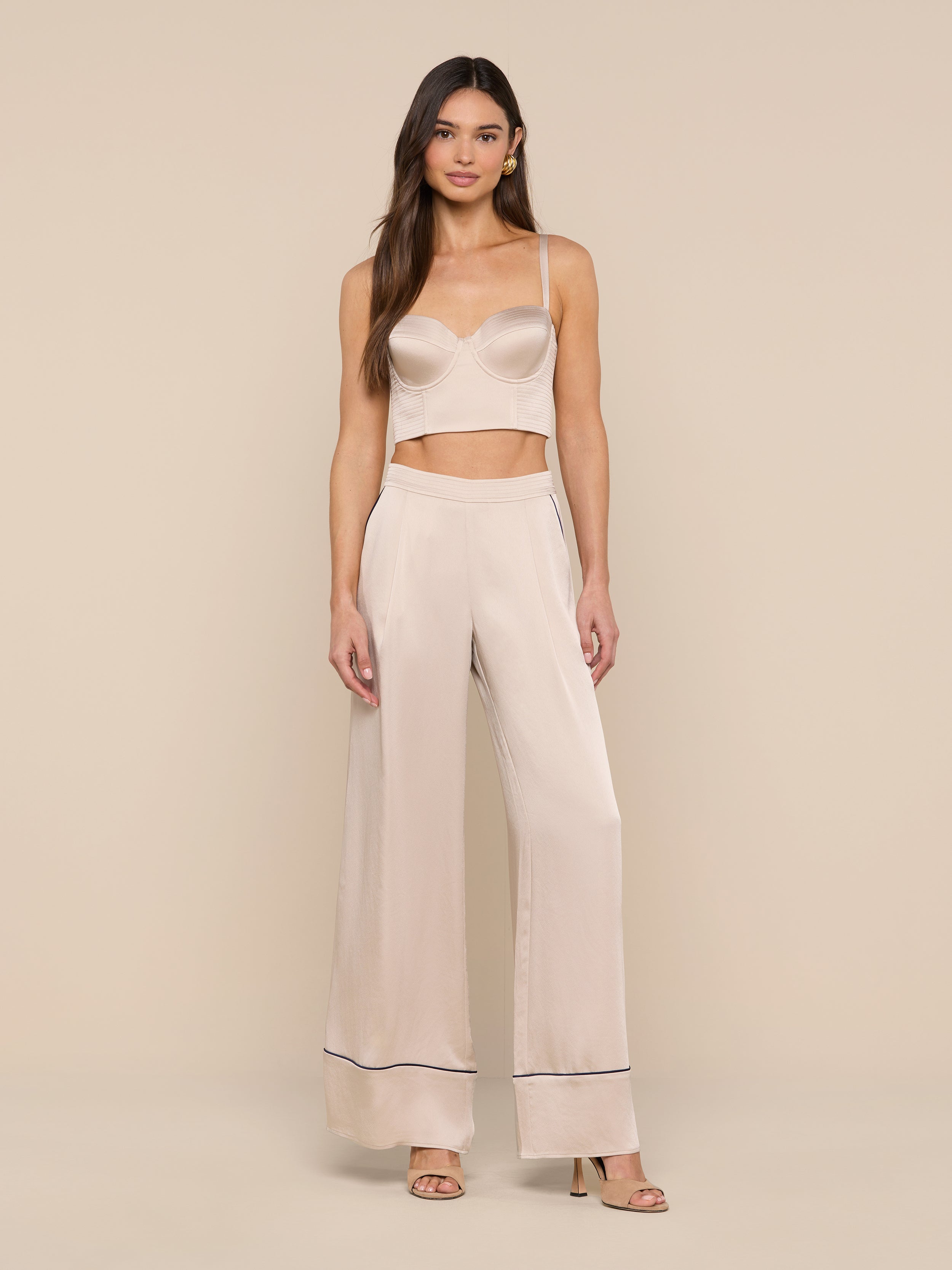 A woman stands against a plain white background wearing a light beige camisole and the Tavia Trouser in matching satin with dark trim at the hems. She has long brown hair and is wearing open-toe heels, capturing the effortless elegance of luxe loungewear.