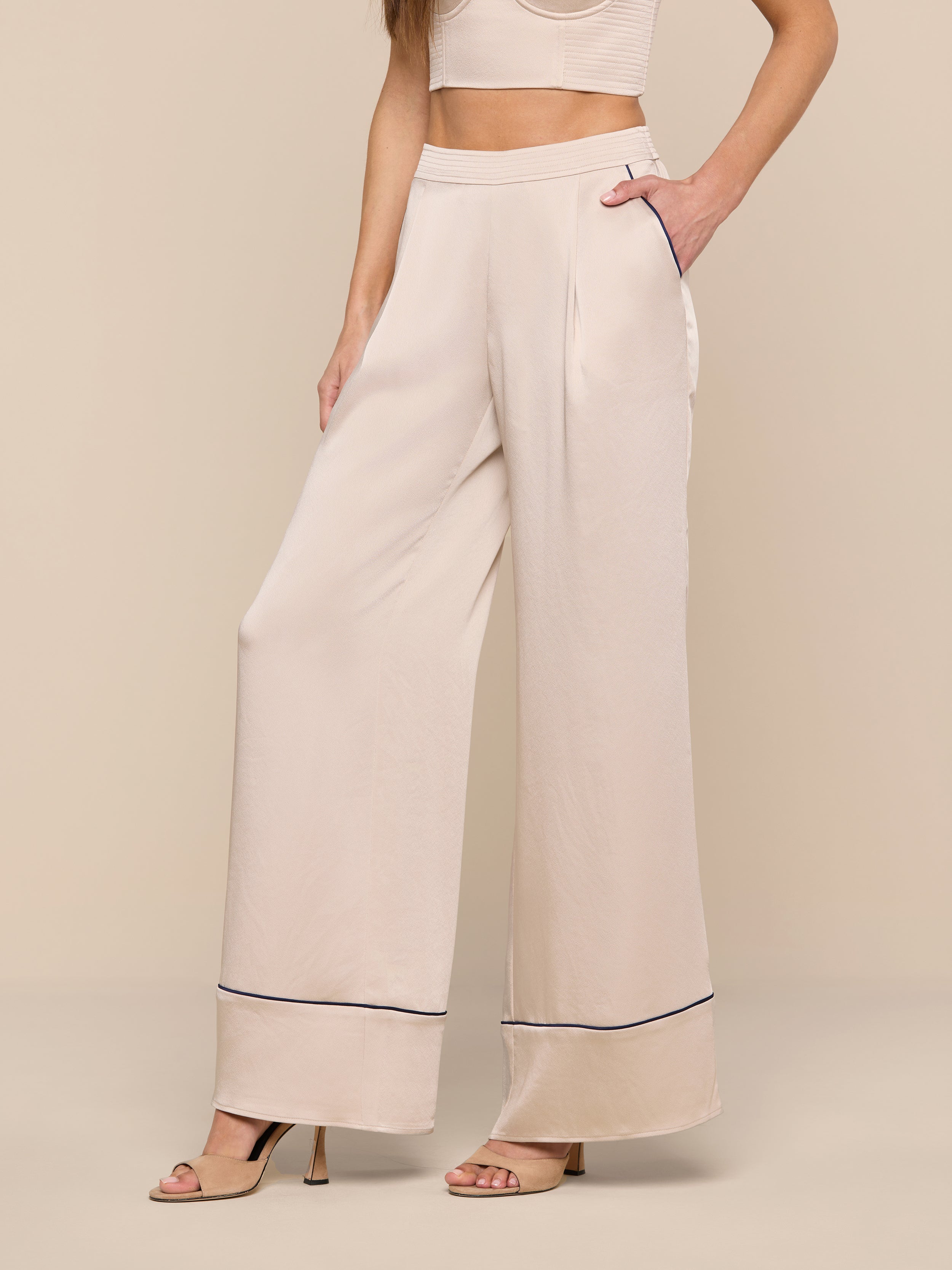 A woman stands against a white background, wearing the high-waisted, wide-leg Tavia Trouser in beige satin featuring black trim near the hem. She pairs it with a matching crop top and nude heeled sandals. One hand rests in her pocket, and her face is not visible.