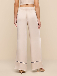 A woman stands facing away, wearing the Tavia Trouser—high-waisted, wide-leg satin trousers with a thin dark trim near the hem—paired with beige heeled sandals and a cropped beige top.