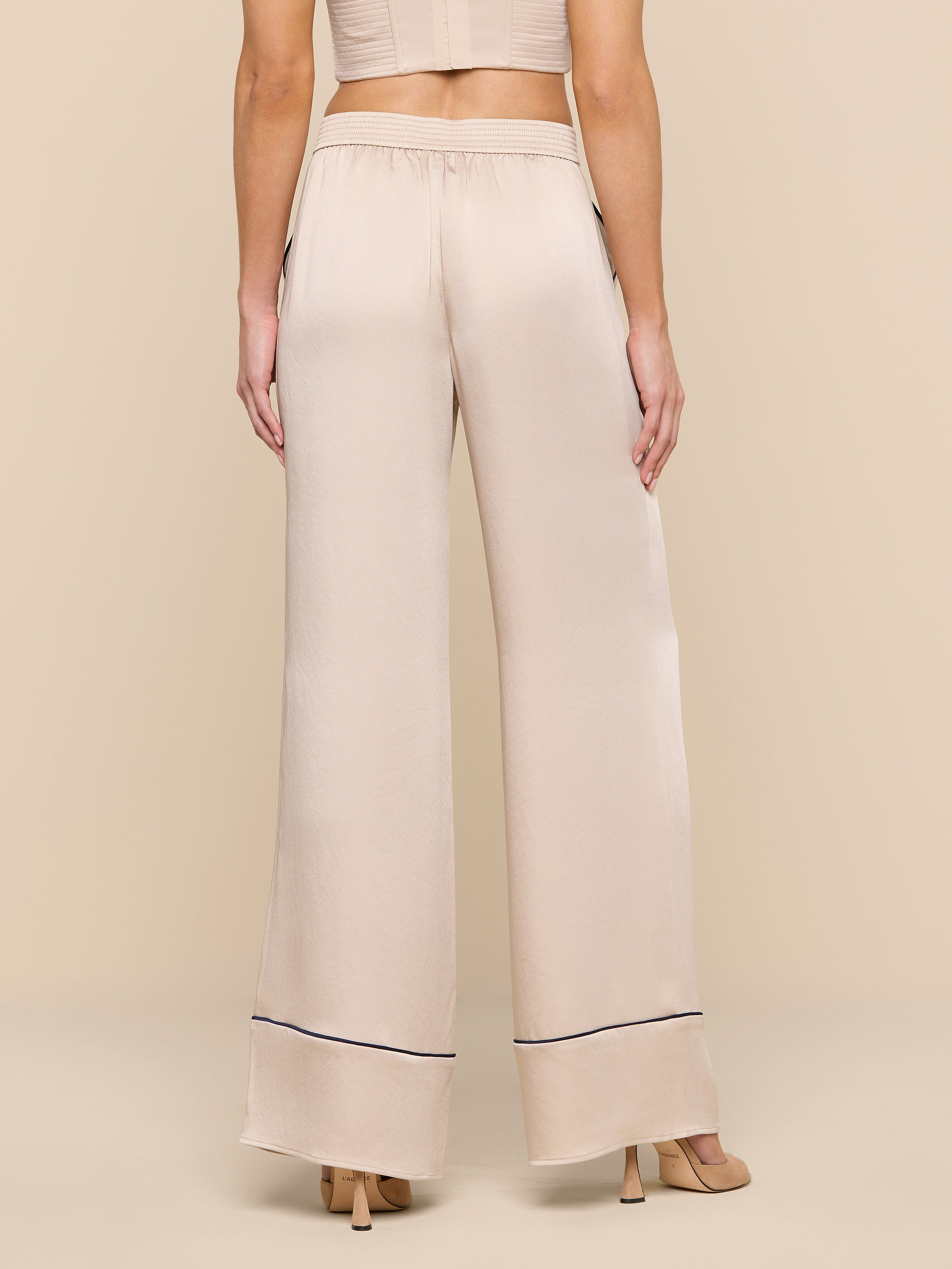 A woman stands facing away, wearing the Tavia Trouser—high-waisted, wide-leg satin trousers with a thin dark trim near the hem—paired with beige heeled sandals and a cropped beige top.