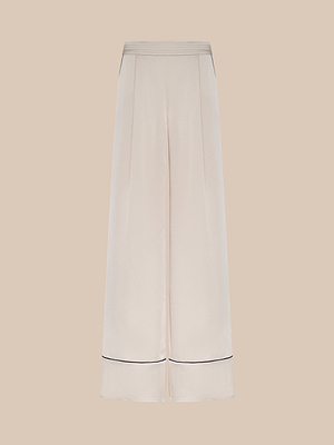 The Tavia Trouser is a wide-leg, high-waisted cream satin pant with subtle pleats, side pockets, and thin black piping near the hem, shown against a beige background.