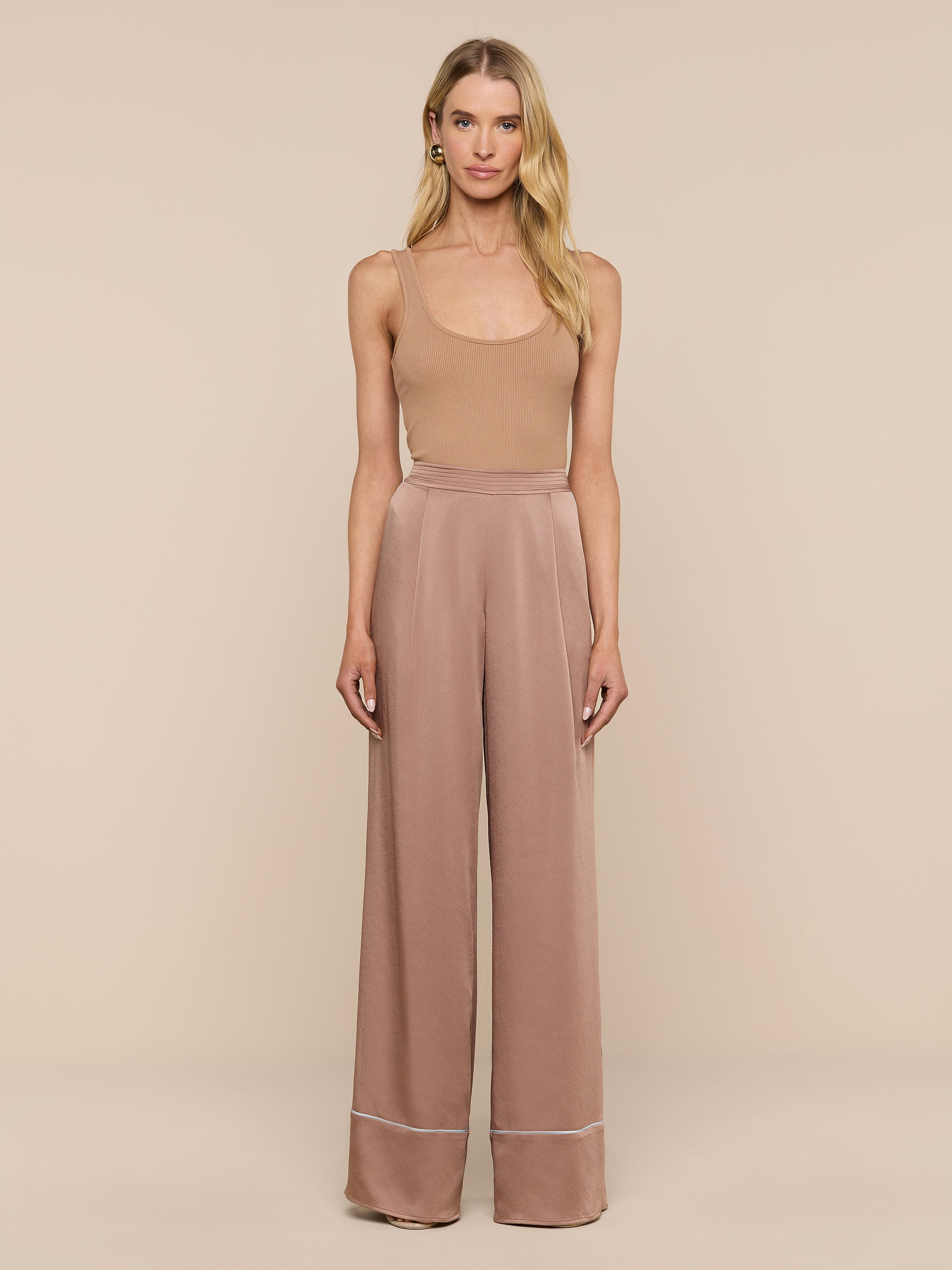 A woman stands facing forward, wearing a tan sleeveless top and high-waisted, wide-leg Tavia Satin Trouser in mauve with white trim, set against a plain white background.