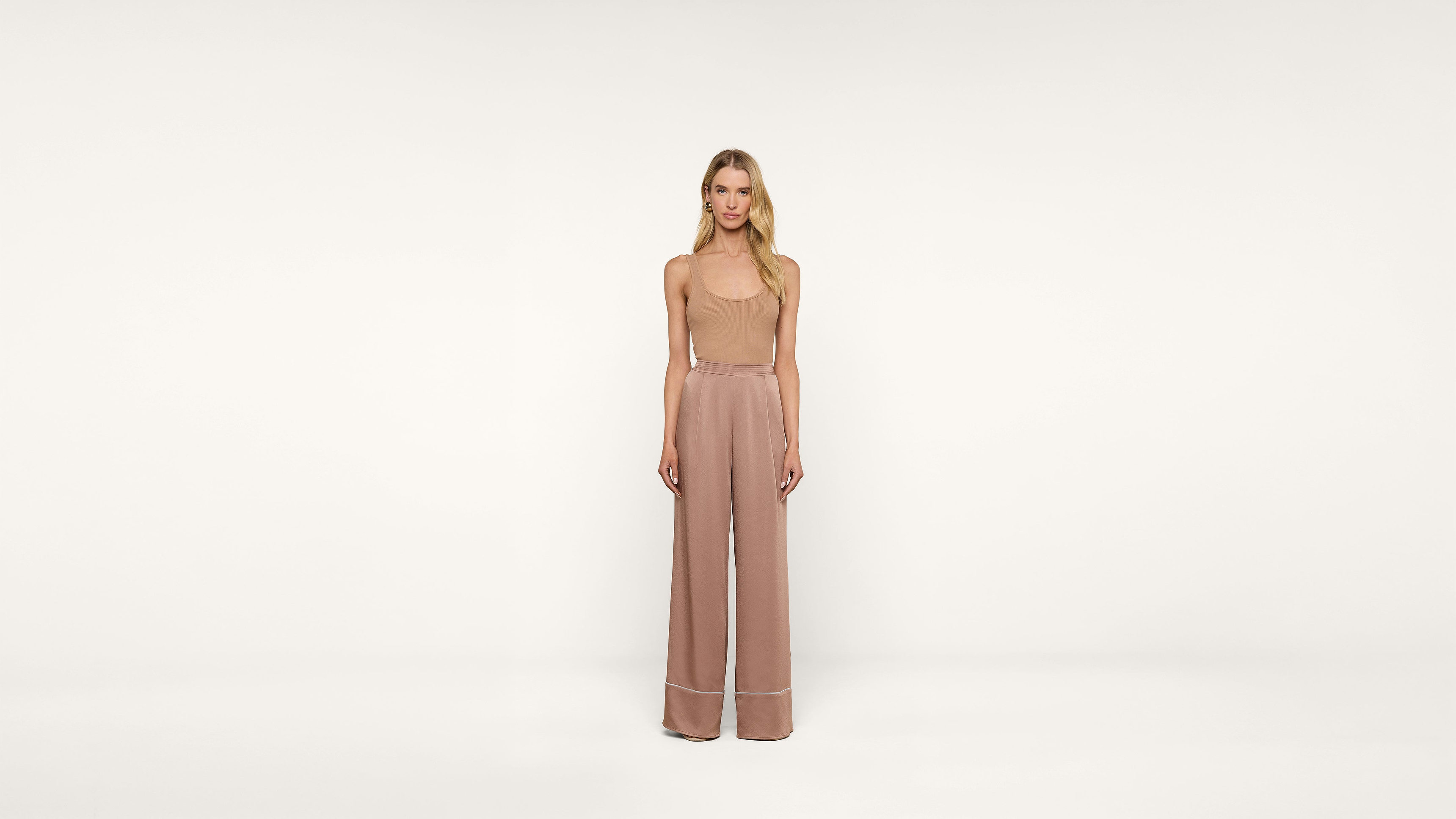 A woman stands facing forward against a plain white background, wearing a sleeveless beige top and high-waisted, loose-fitting, wide-legged light brown pants.