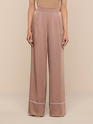 A person stands against a white background wearing the chic Tavia Satin Trouser—high-waisted and wide-leg in a light brown, subtly lustrous fabric, finished with a thin white stripe near the hem. The upper body is partially visible.