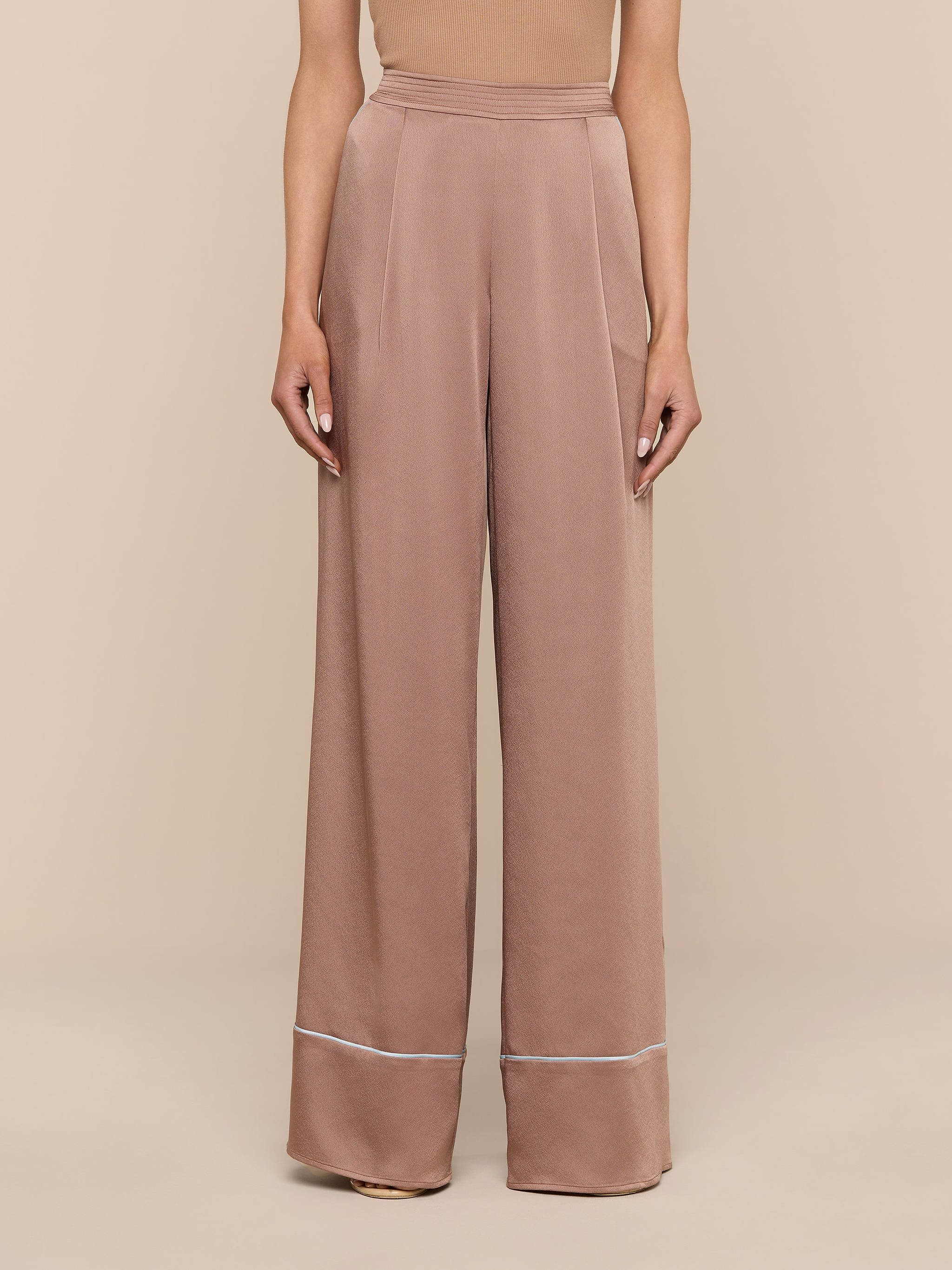 A person stands against a white background wearing the chic Tavia Satin Trouser—high-waisted and wide-leg in a light brown, subtly lustrous fabric, finished with a thin white stripe near the hem. The upper body is partially visible.