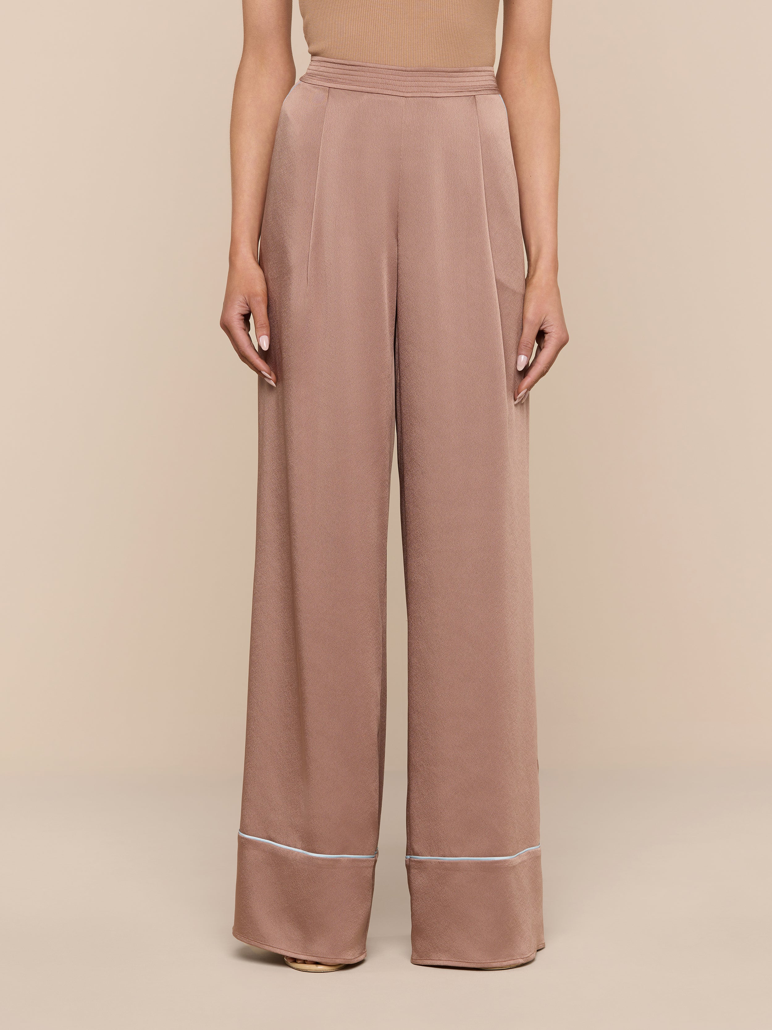 A person stands against a white background wearing the chic Tavia Satin Trouser—high-waisted and wide-leg in a light brown, subtly lustrous fabric, finished with a thin white stripe near the hem. The upper body is partially visible.