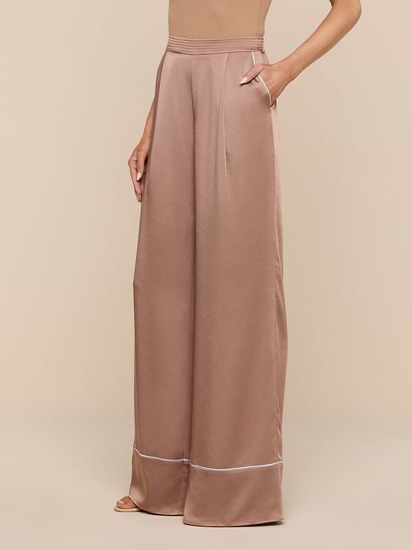 A person models the Tavia Satin Trouser—a pair of high-waisted, wide-leg rose gold satin pants with pockets—against a plain white background, showing only the lower torso, legs, and one arm.