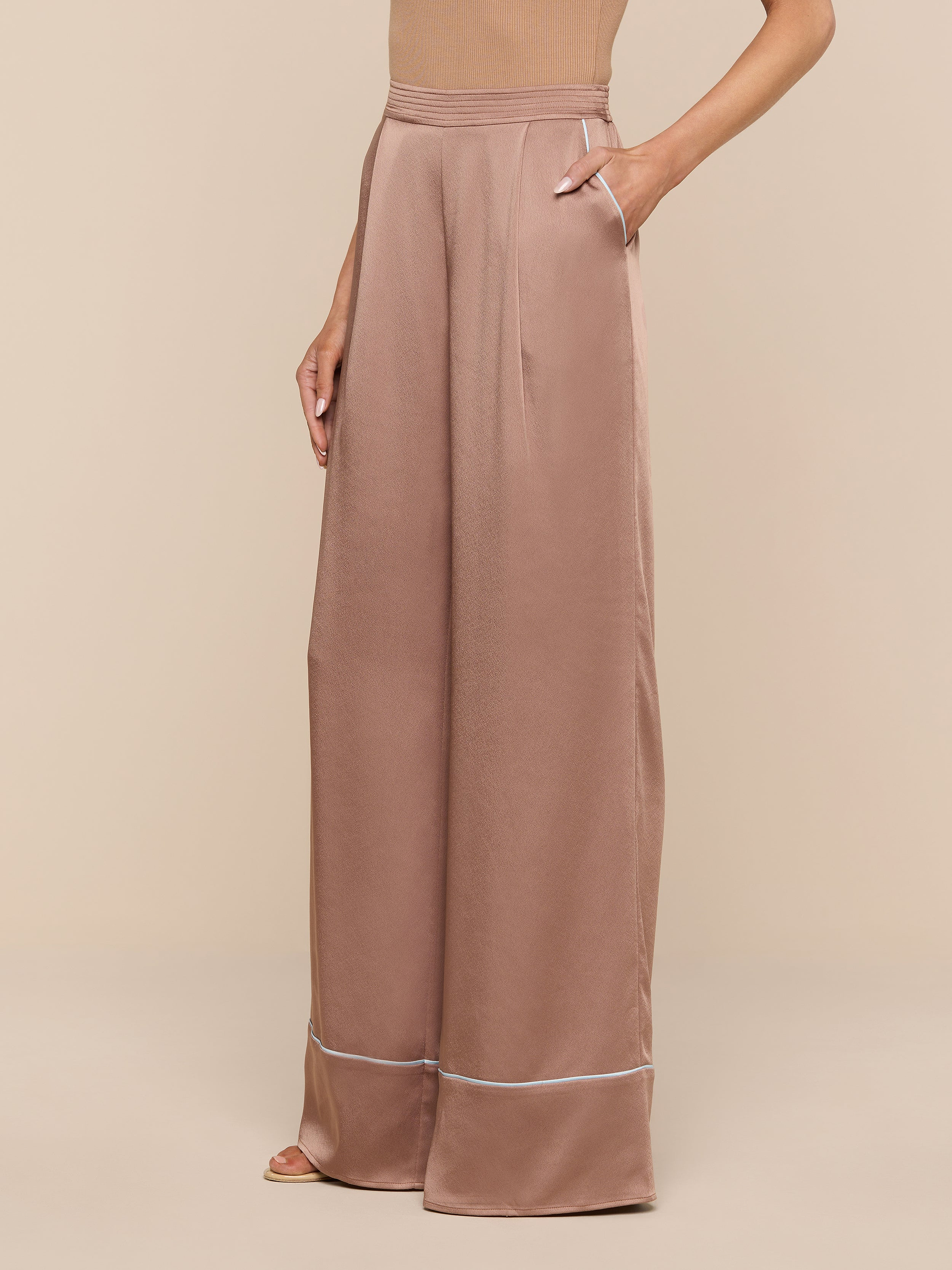 A person models the Tavia Satin Trouser—a pair of high-waisted, wide-leg rose gold satin pants with pockets—against a plain white background, showing only the lower torso, legs, and one arm.