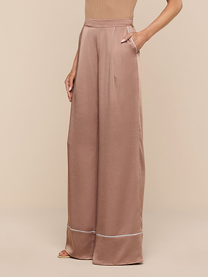 A person wears the Tavia Satin Trouser in dusty rose, featuring a high waist, wide-leg cut, subtle sheen, and white contrast piping near the hem. Their left hand is in the pocket against a plain beige background.