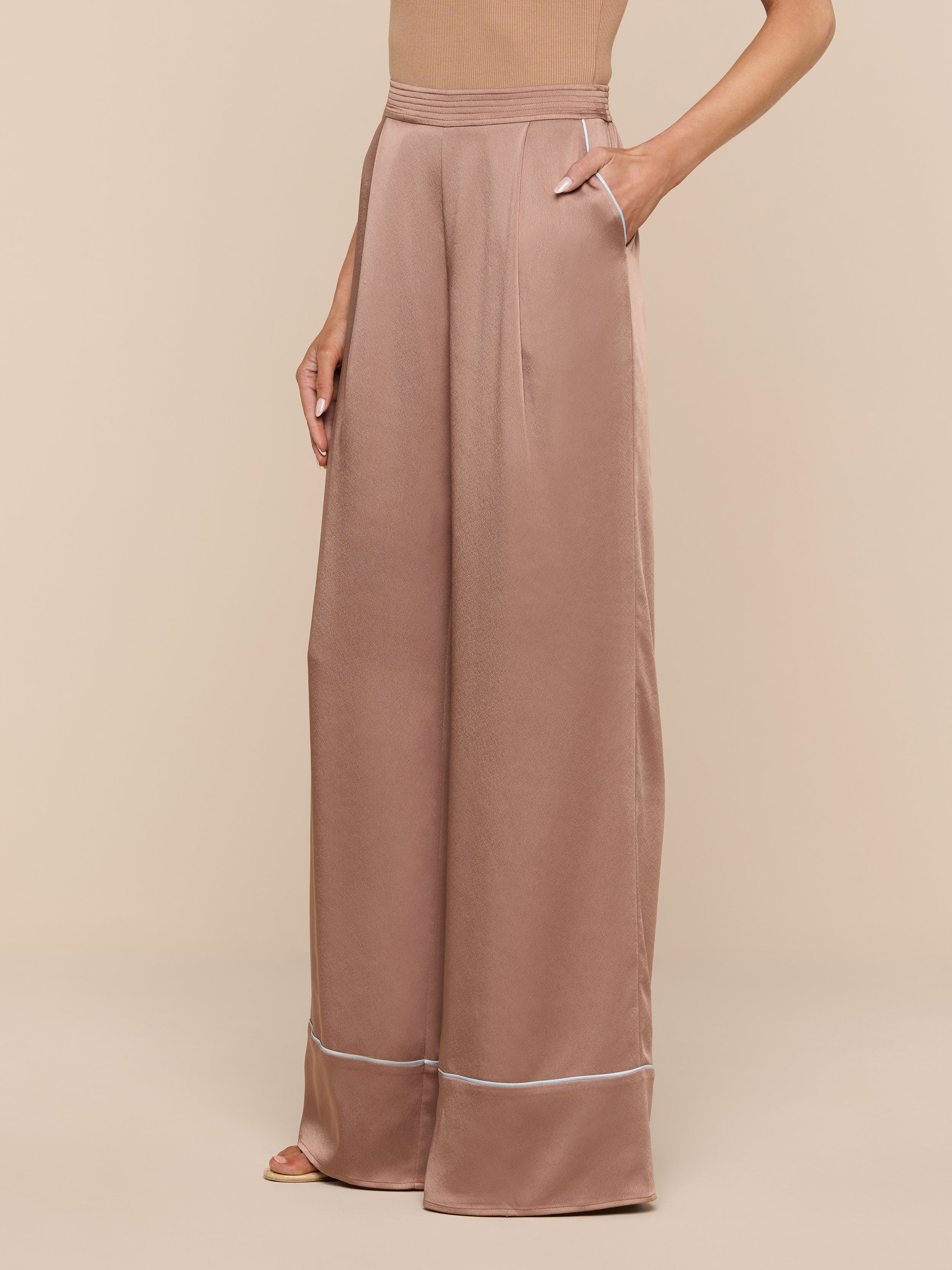 A person models the Tavia Satin Trouser—a pair of high-waisted, wide-leg rose gold satin pants with pockets—against a plain white background, showing only the lower torso, legs, and one arm.