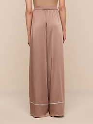 A person shown from the back against a plain background wears the Tavia Satin Trouser—chic, high-waisted, wide-leg pants in light brown satin, featuring a white stripe near the hem.