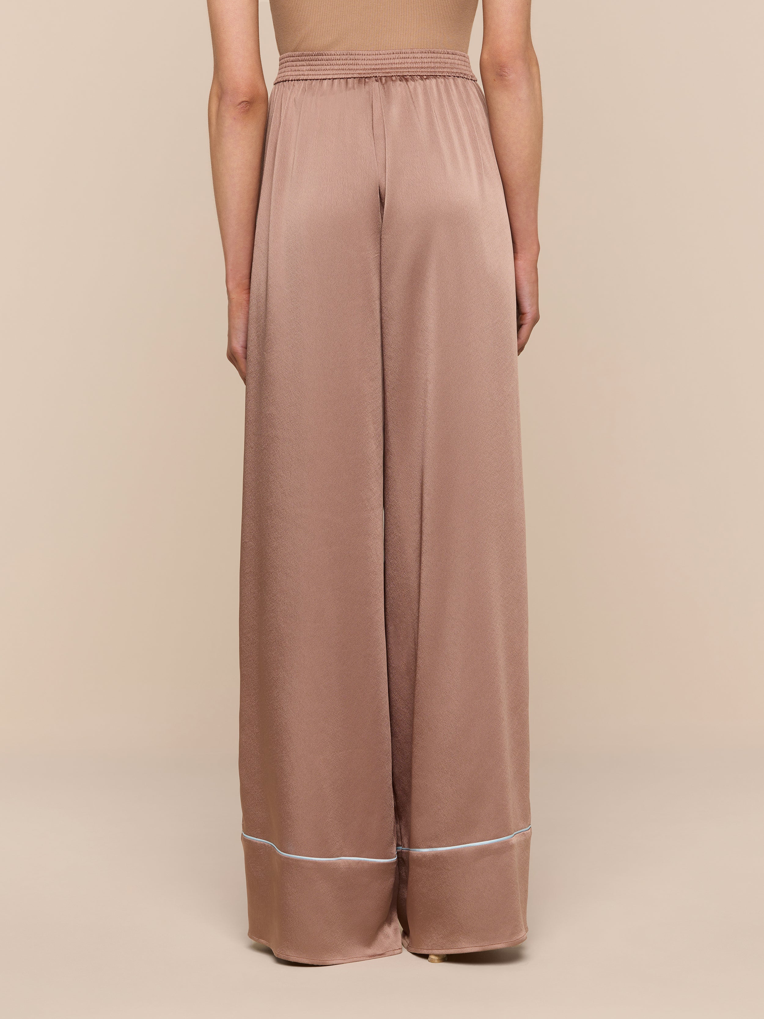 A person shown from the back against a plain background wears the Tavia Satin Trouser—chic, high-waisted, wide-leg pants in light brown satin, featuring a white stripe near the hem.