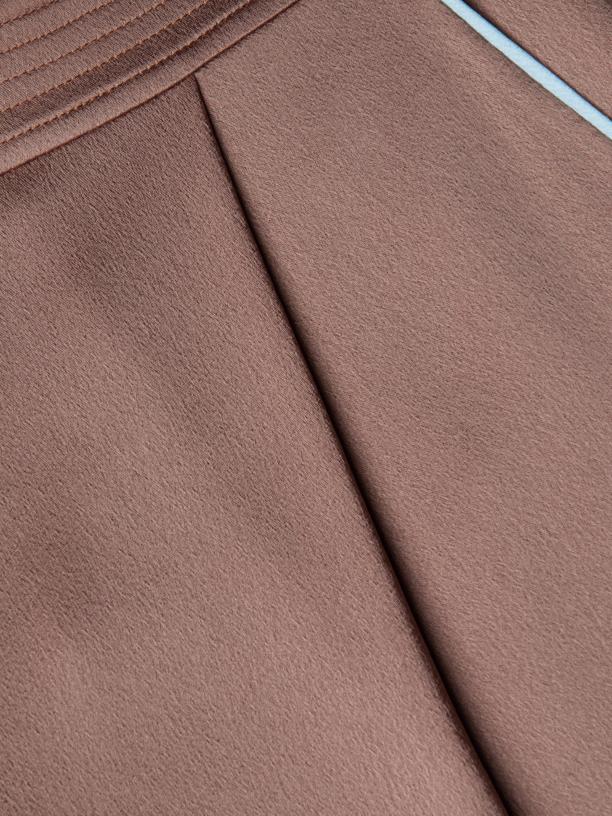 Close-up of a brown textured satin fabric with pleats and a diagonal seam, featuring light blue trim along the edge, as seen on the chic Tavia Satin Trouser.